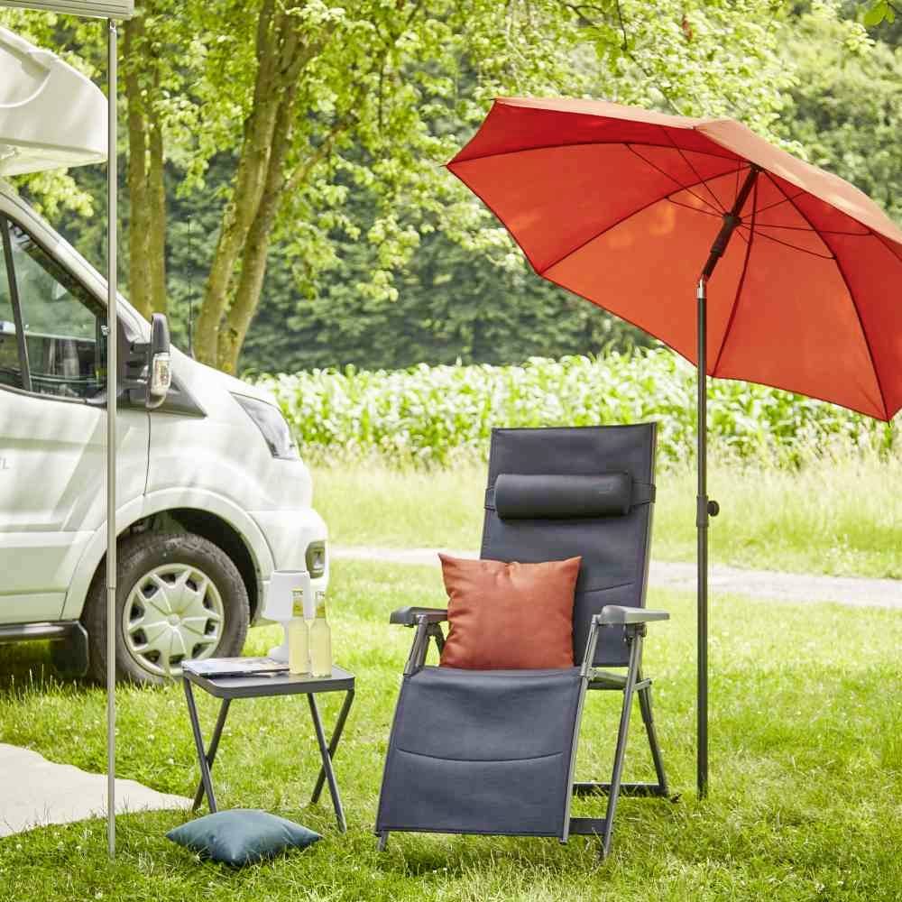 Camping-car garé dans un environnement verdoyant avec une table, un transat et un parasol rouge. Au fond, une prairie et des arbres, des bouteilles de boisson sur la table.