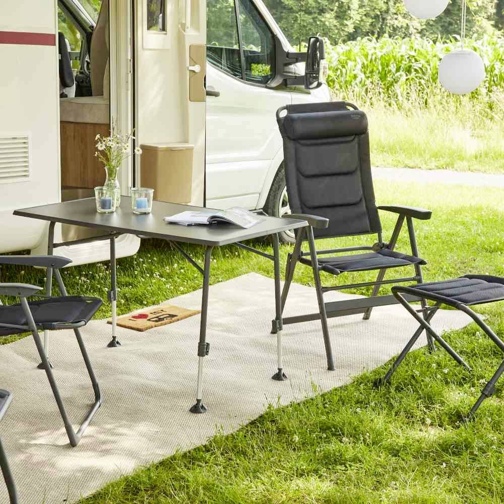 Camping-car avec porte ouverte, deux chaises et une table sur un tapis dans un environnement verdoyant. Des lanternes lumineuses suspendues. Ambiance extérieure détendue.