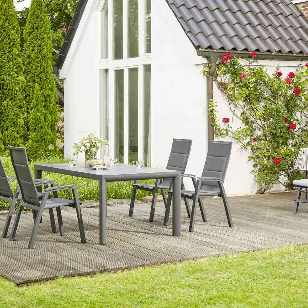 Un jardin moderne avec un sol en bois, une table à manger et des chaises devant une petite maison ; des rosiers grimpants le long du mur blanc de la maison.