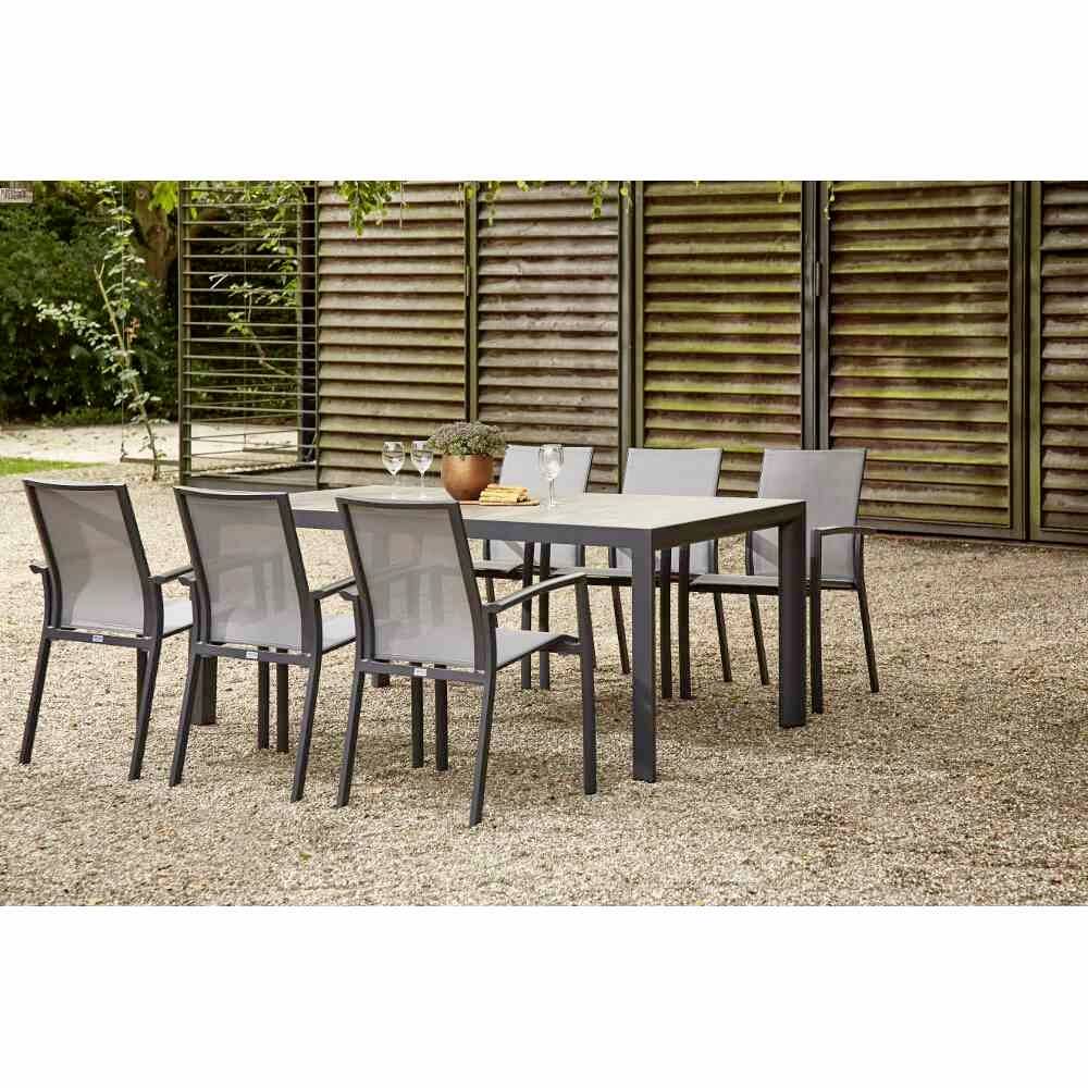 Une table de jardin avec six chaises sur un sol de gravier ; derrière, des murs en bois et des plantes. Sur la table, un pot de fleurs et deux verres.