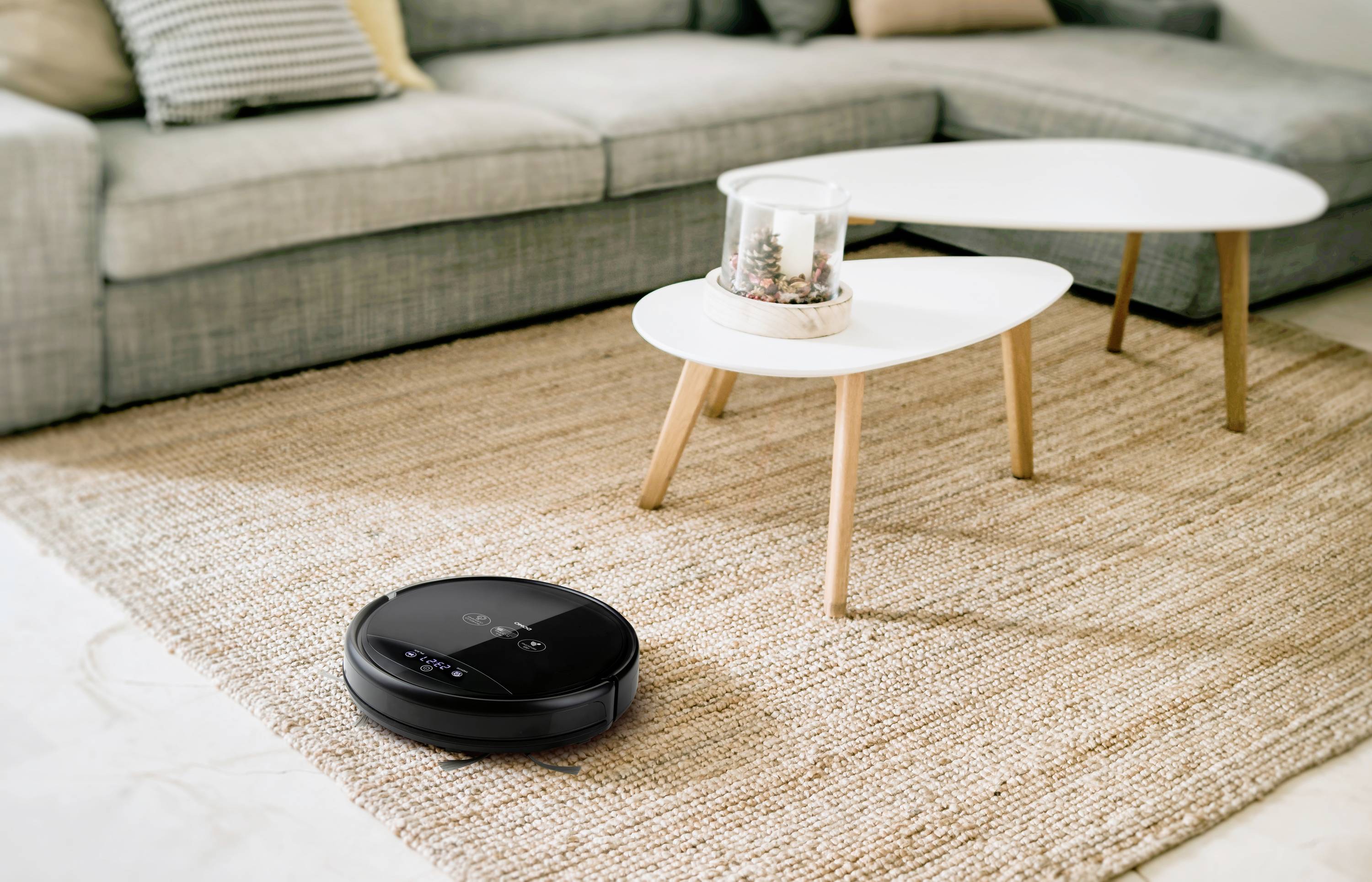 Un robot aspirateur noir nettoie un tapis beige-brun dans un salon avec un canapé gris et des tables d'appoint blanches.