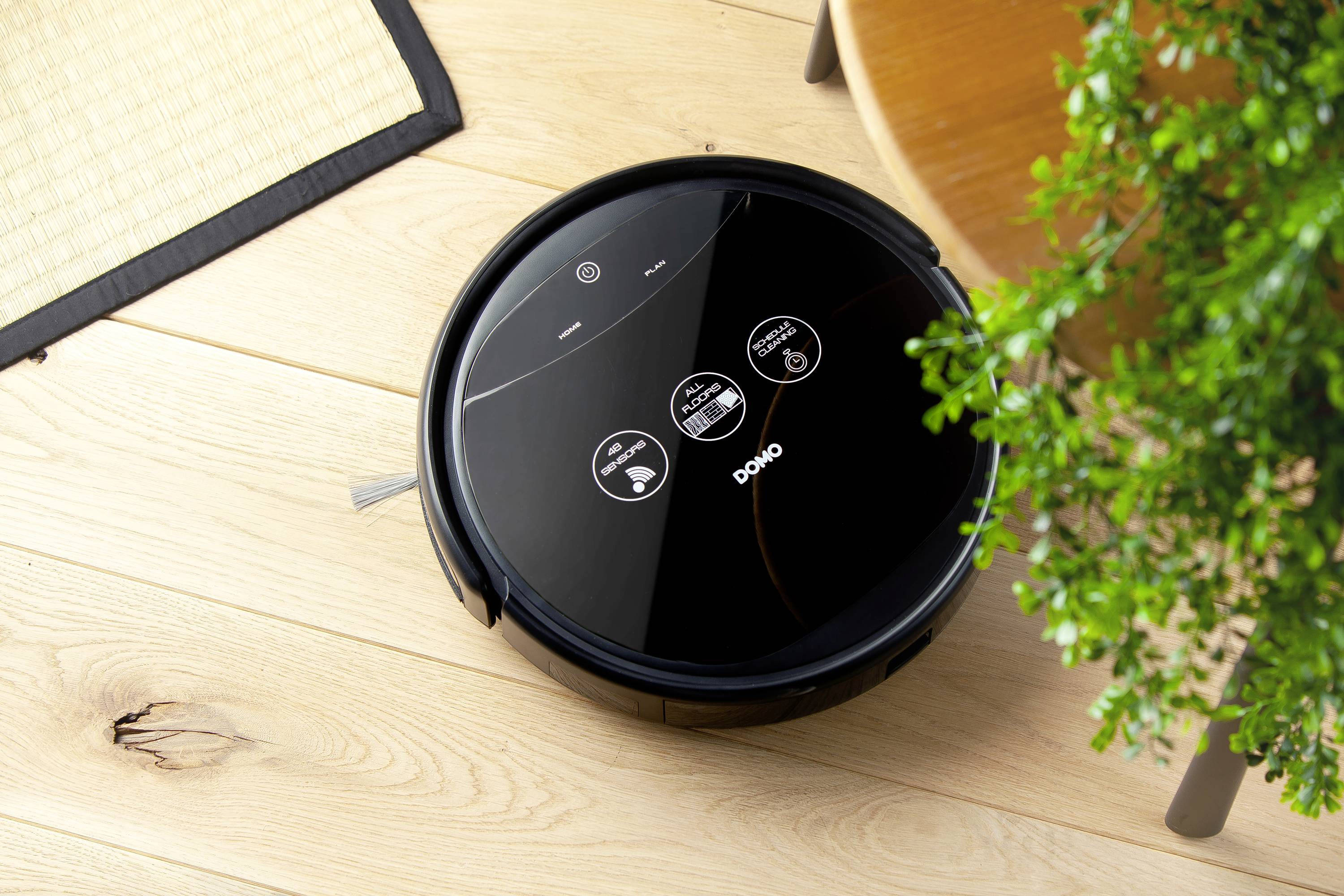 Un robot aspirateur noir et rond avec un panneau de commande tactile sur un parquet, à côté d'une table et d'une plante verte.