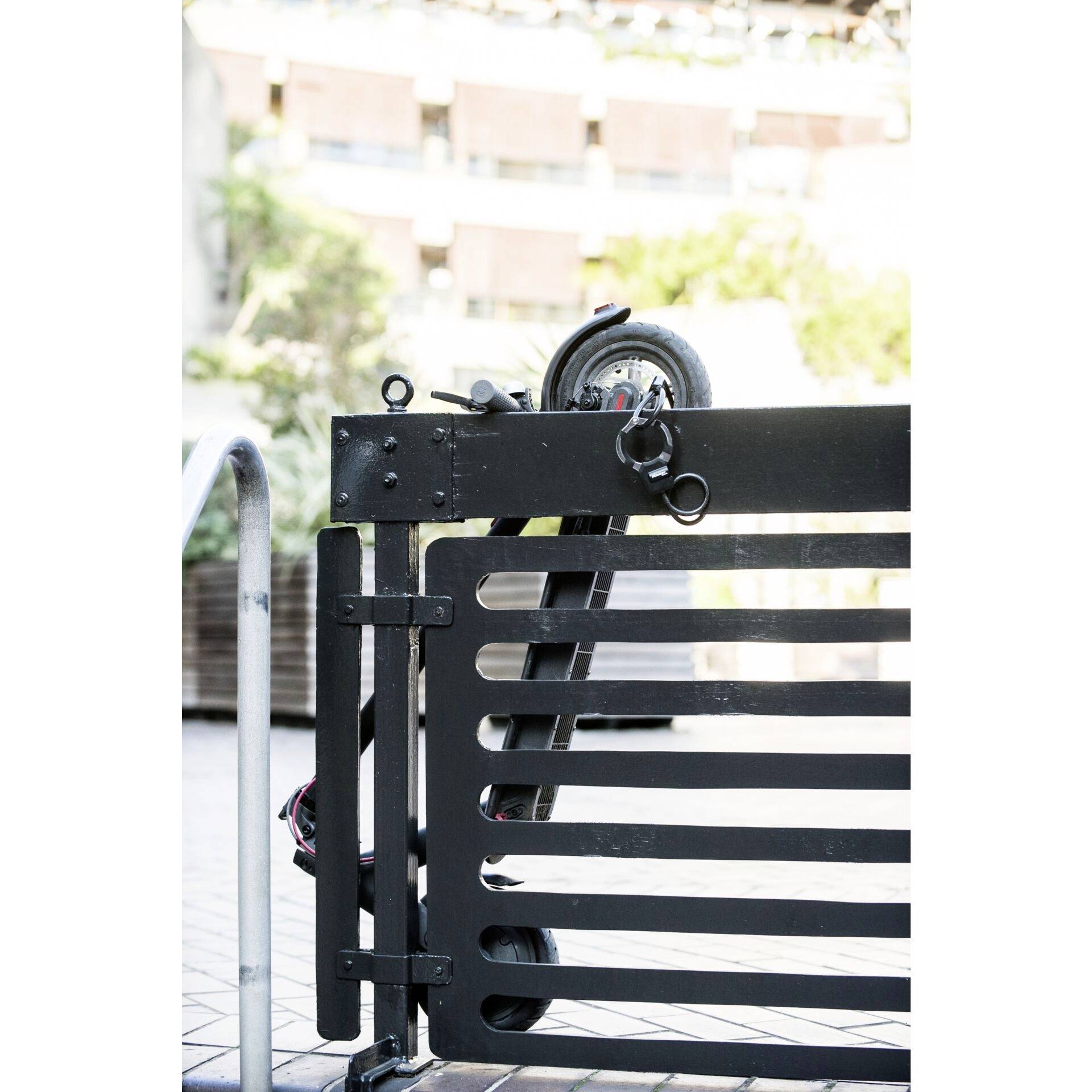 Une grille métallique noire avec un verrou à charnière, un bâtiment flou et des plantes vertes visibles en arrière-plan.