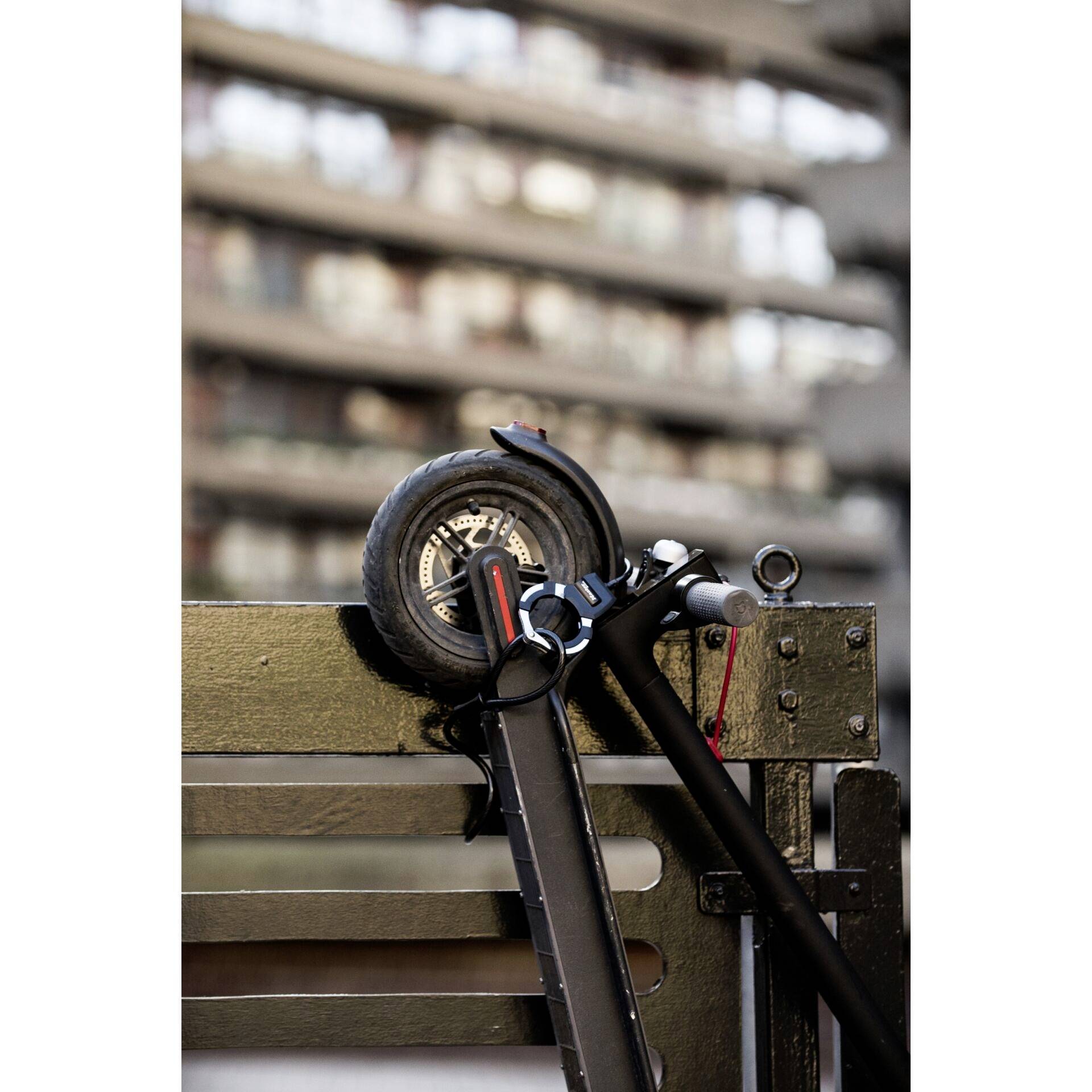 Un scooter noir est appuyé contre une rambarde métallique. L'arrière-plan est flou et montre un bâtiment à plusieurs étages.