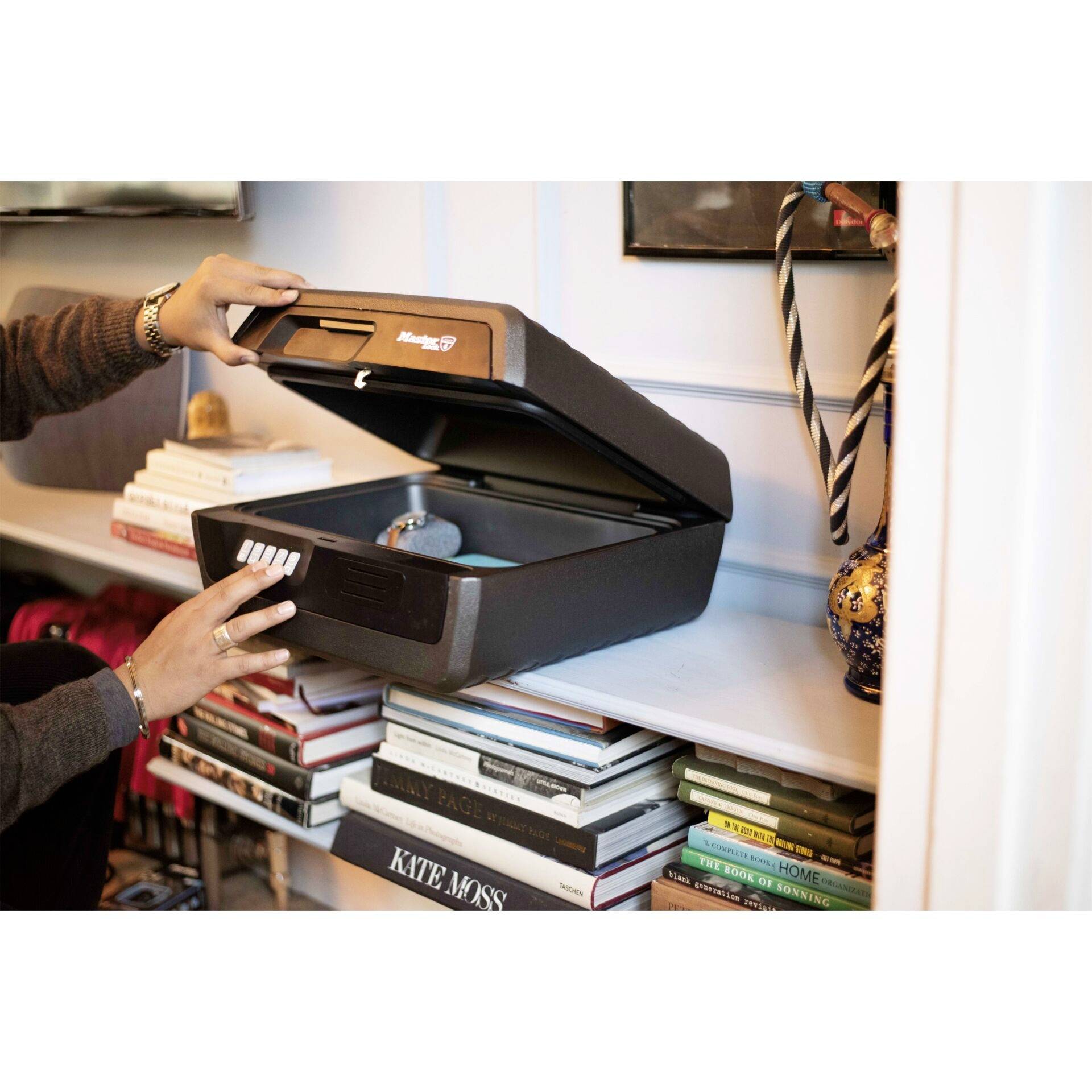 Une personne ouvre un coffre-fort avec une main sur une étagère de bibliothèque. Le coffre-fort est noir et contient des objets.