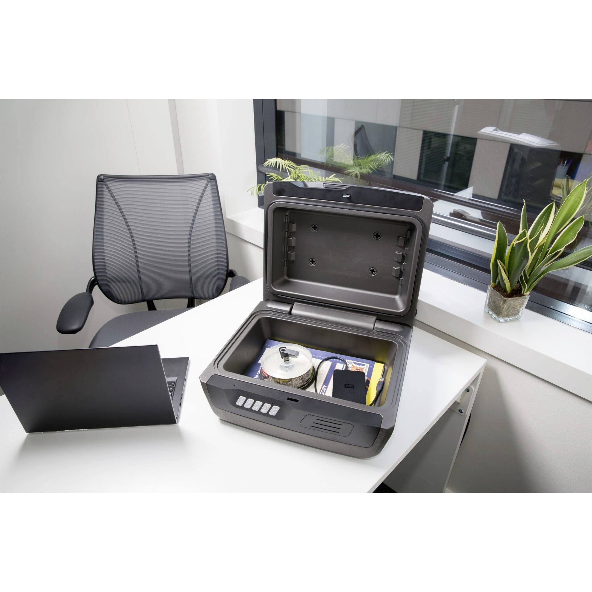 Dans le bureau, un coffre-fort ouvert est posé sur un bureau. À l'intérieur du coffre-fort se trouvent des CD et des documents. Un ordinateur portable et une chaise de bureau sont à côté.