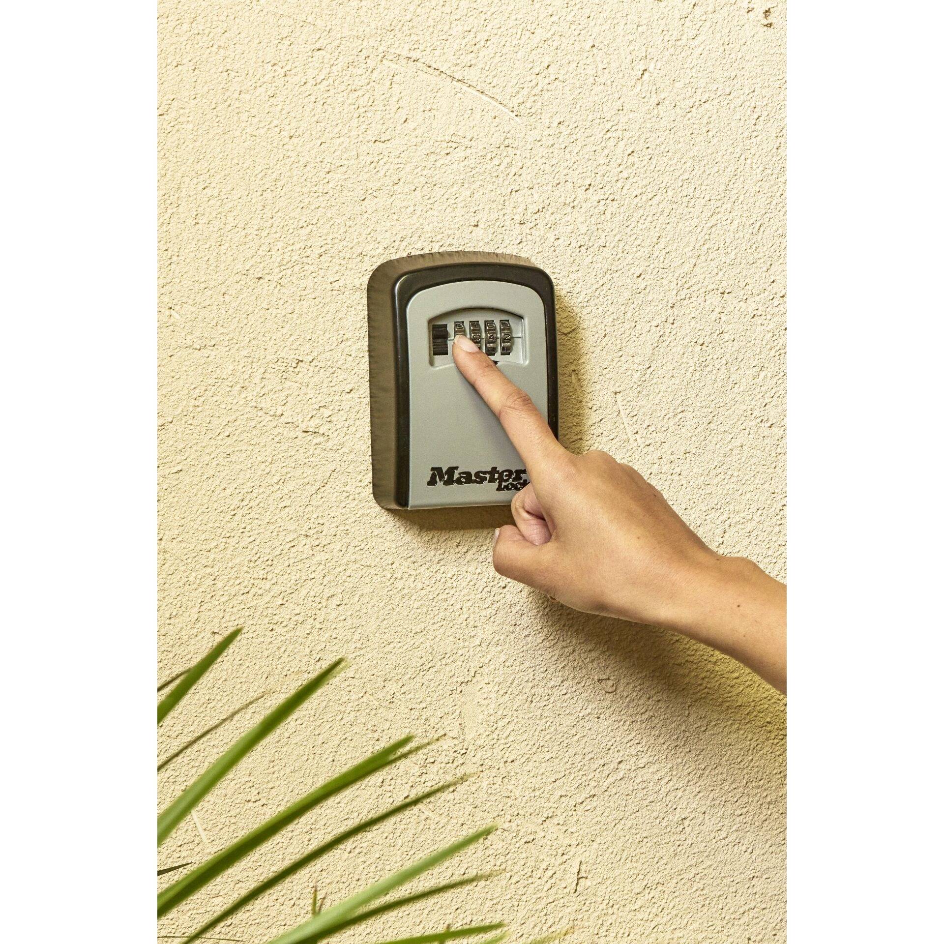 Une main entre une combinaison dans un cadenas à chiffres sur un mur pour ouvrir un coffre. En dessous se trouve l'inscription « Master ».