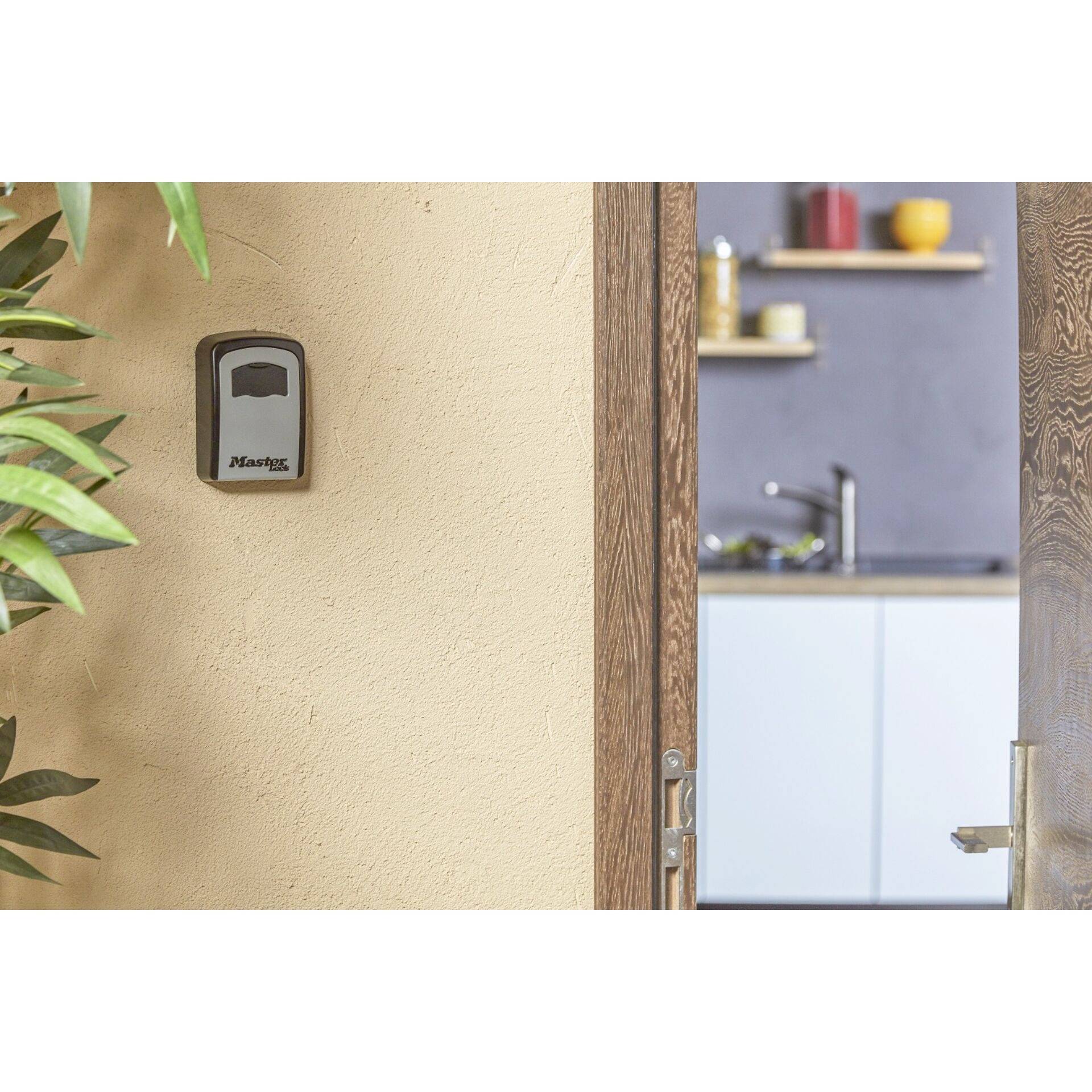 Une scène de cuisine vue à travers une porte en bois ouverte, montrant un coffre-fort à clés accroché au mur et des étagères avec de la vaisselle et des épices en arrière-plan.