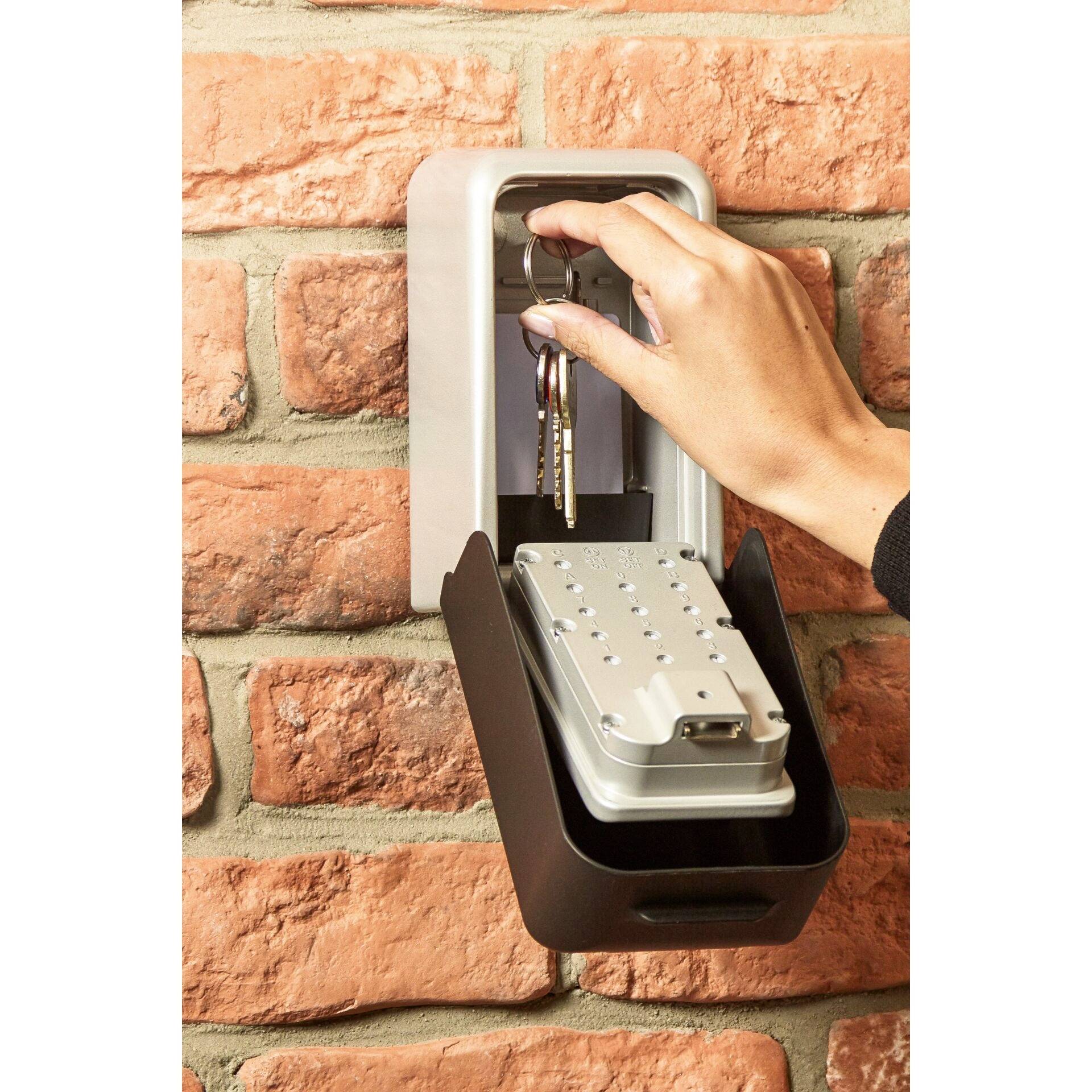 Une main prend un trousseau de clés dans un coffre-fort à clés ouvert sur un mur de briques. Le coffre-fort est rectangulaire et gris foncé.