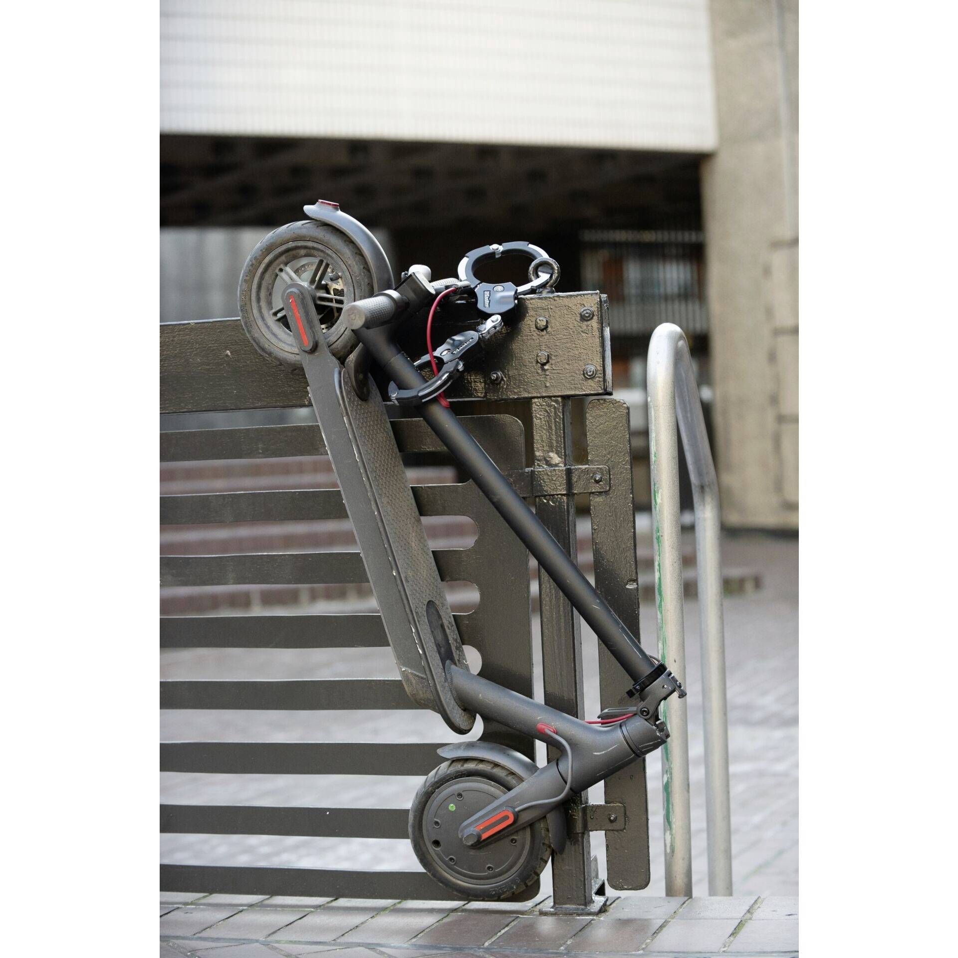 Une trottinette électrique est sécurisée avec un antivol et est posée en diagonale contre un portail métallique sur un trottoir dans un environnement urbain.
