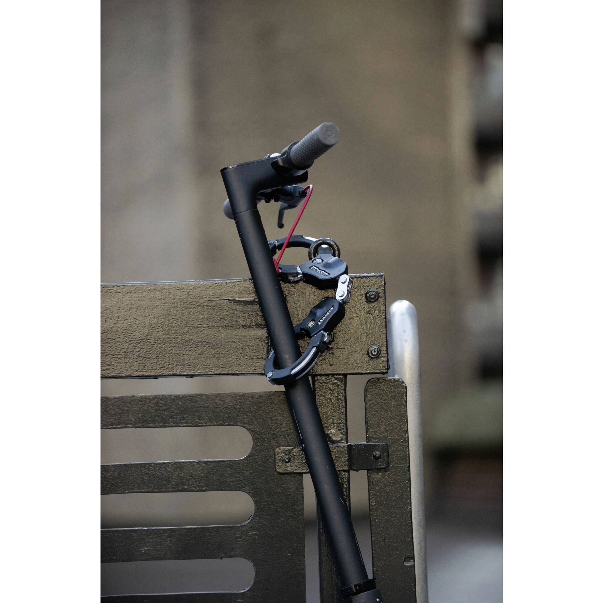 Un trottinette électrique noir est sécurisé avec un cadenas sur une clôture métallique. L'arrière-plan est flou.