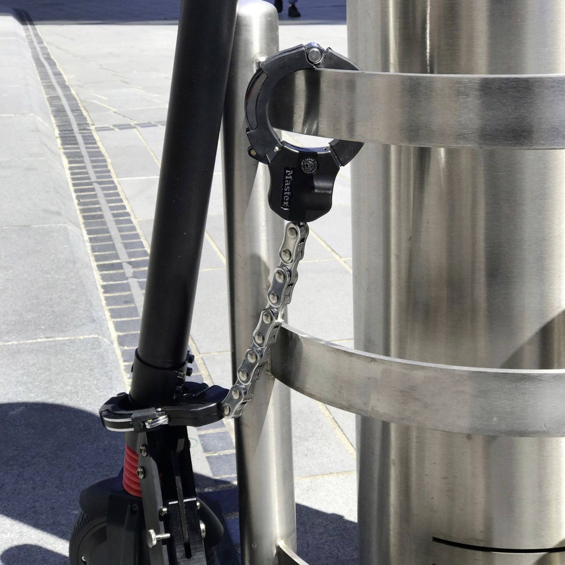 Une trottinette électrique est attachée à un poteau métallique avec une chaîne en acier. L'environnement semble être urbain.