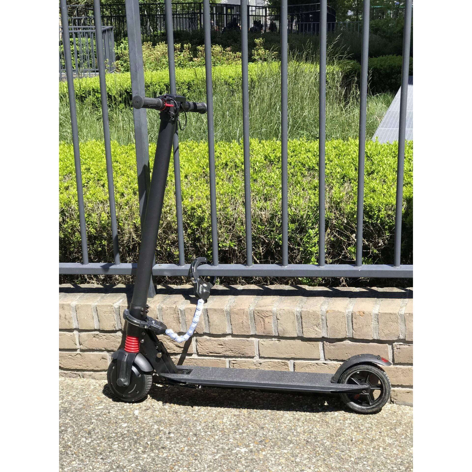 Un scooter électrique noir est stationné devant une clôture métallique sur un chemin pavé. Des plantes vertes sont visibles en arrière-plan.