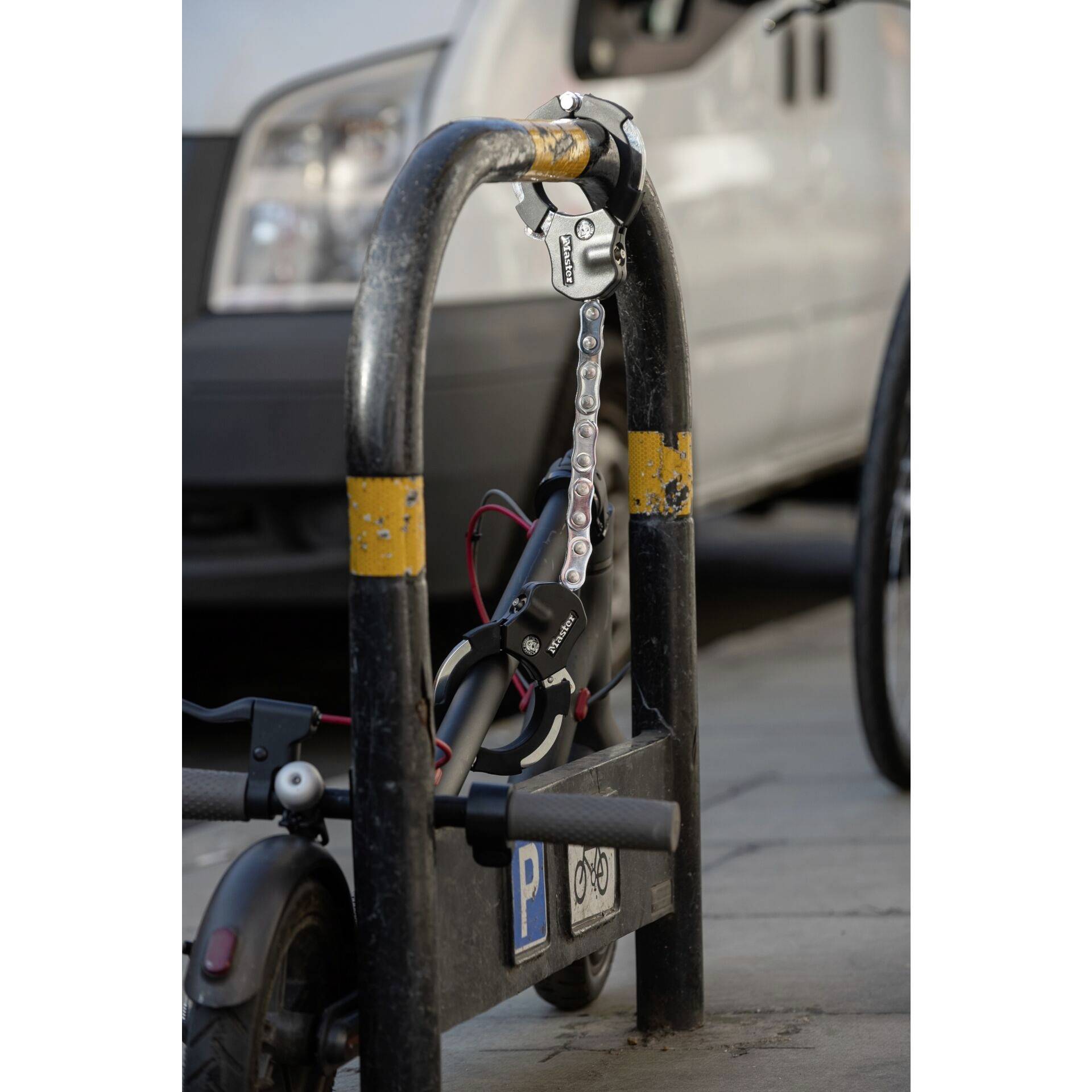 Un scooter électrique sécurisé avec un cadenas à chaîne sur un support noir. Une voiture est visible en arrière-plan.