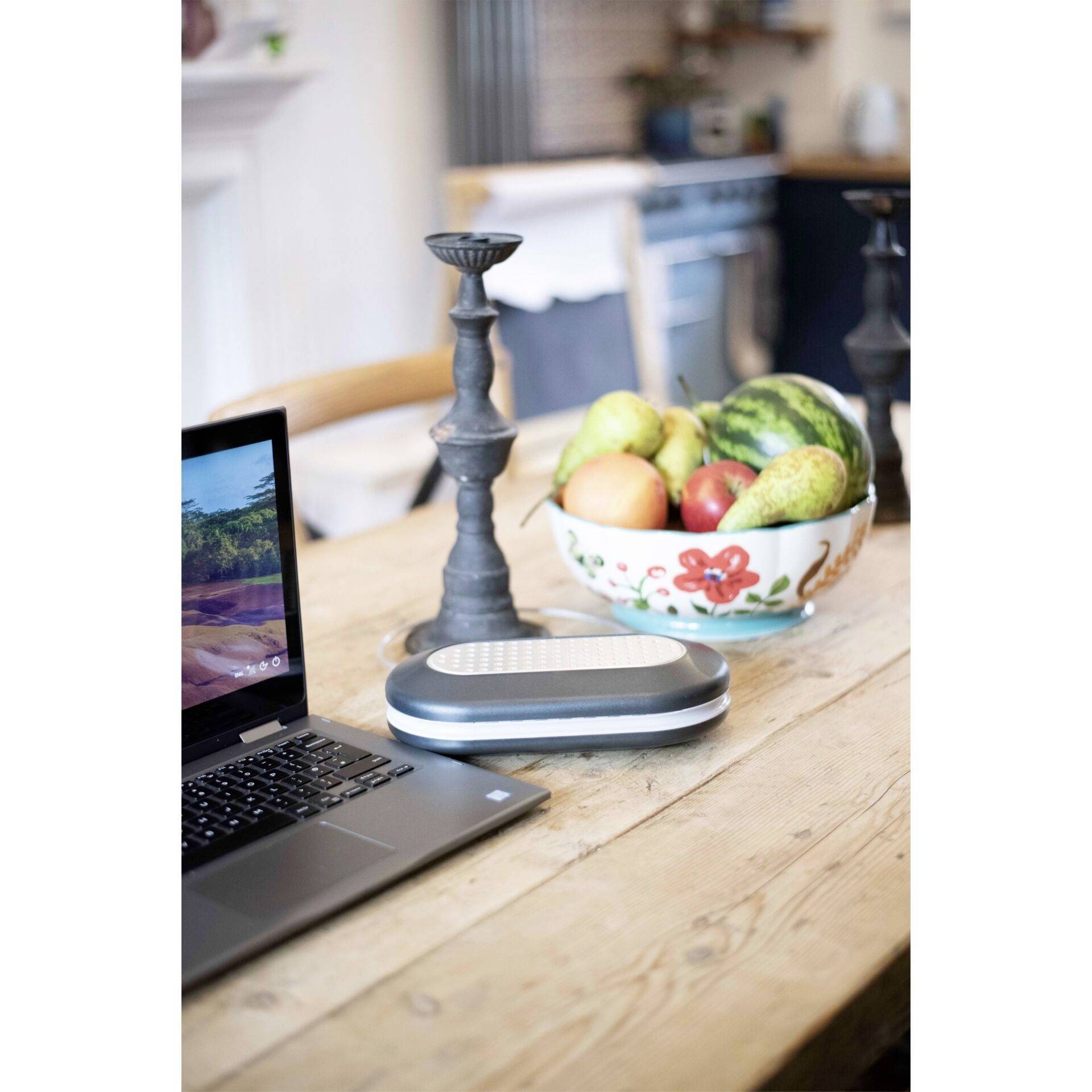 Un ordinateur portable et un haut-parleur sont posés sur une table en bois. En arrière-plan, on peut voir un bol de fruits, dont des melons.