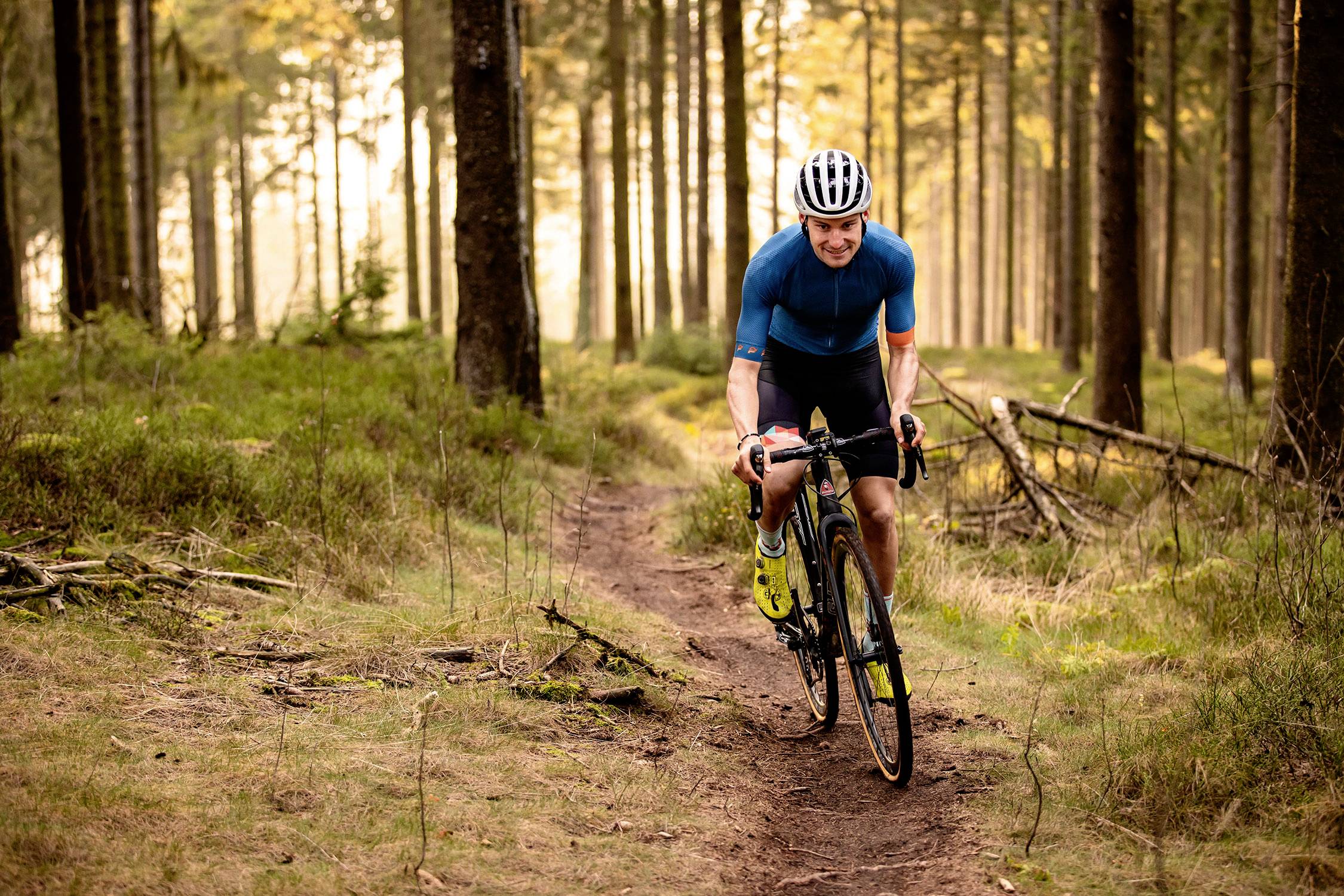 Un cycliste portant un maillot bleu roule sur un sentier étroit à travers une forêt dense, inondée de lumière solaire.