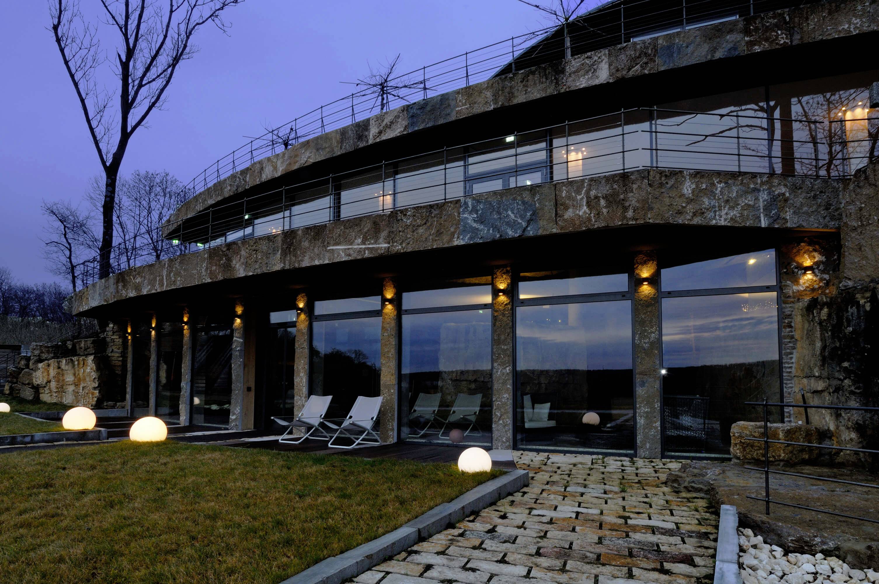 Un bâtiment moderne avec une façade vitrée et des éléments de pierre ronds au crépuscule. Au premier plan, des chemins pavés et des boules lumineuses sont disposés.