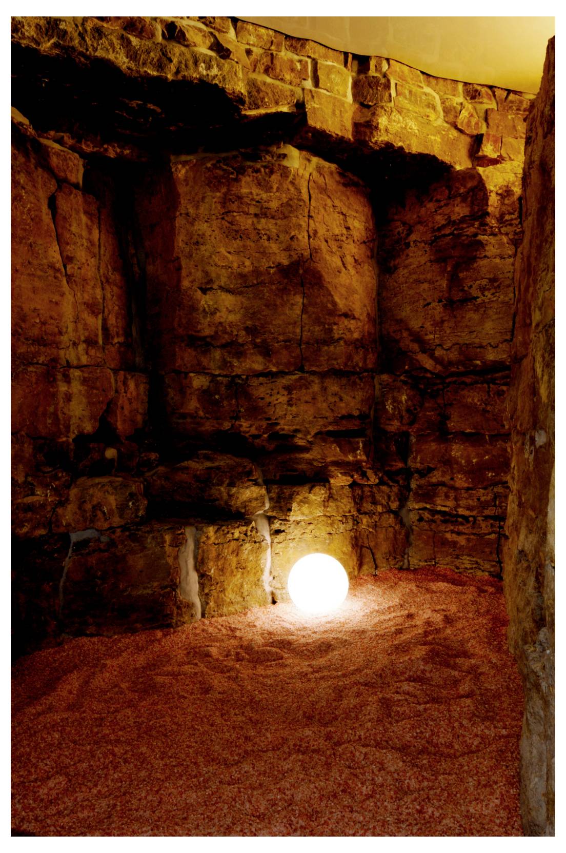 Grotte de pierre sombre aux parois et au sol de roche brute, éclairée par une sphère lumineuse brillante près du fond, au centre.