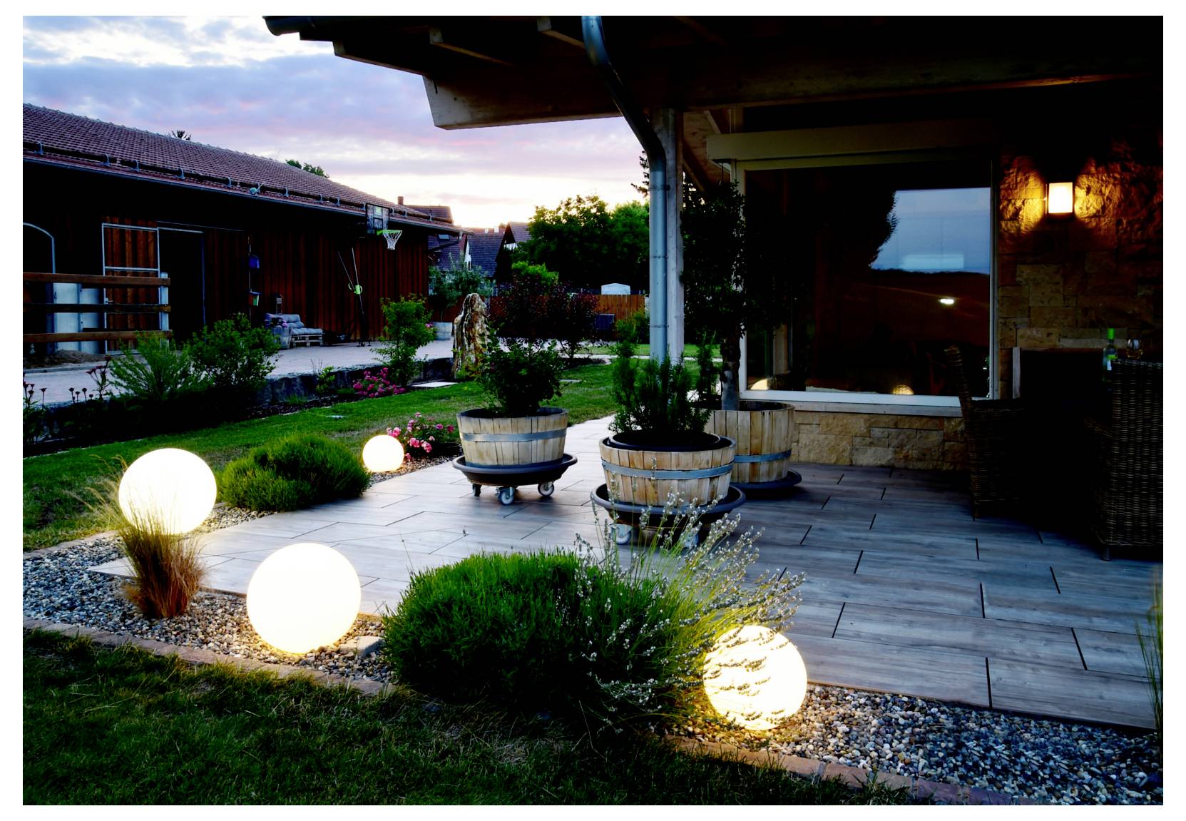 Terrasse et jardin en soirée avec des lumières sphériques lumineuses, des plantes en pot et un dallage en pierre à côté d'une maison au coucher du soleil.