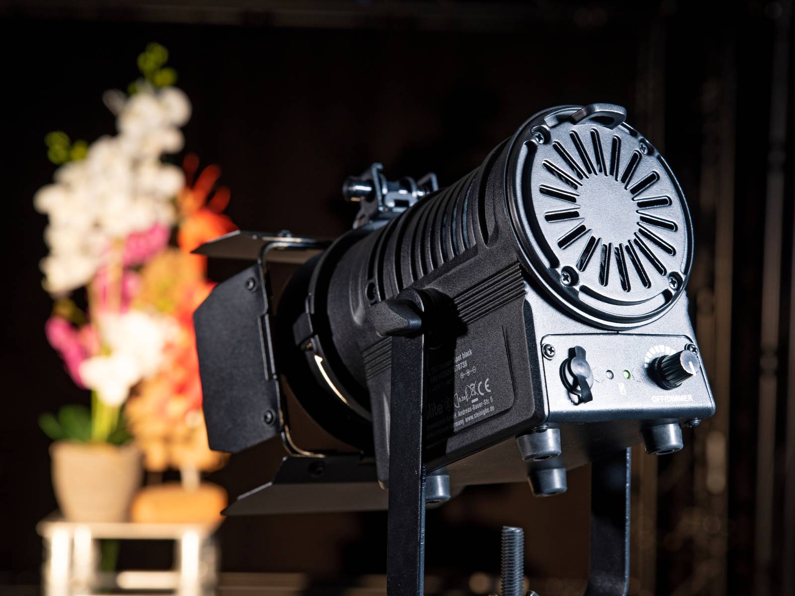 Un projecteur de scène est braqué sur une décoration florale multicolore en arrière-plan. La scène suggère une représentation théâtrale.