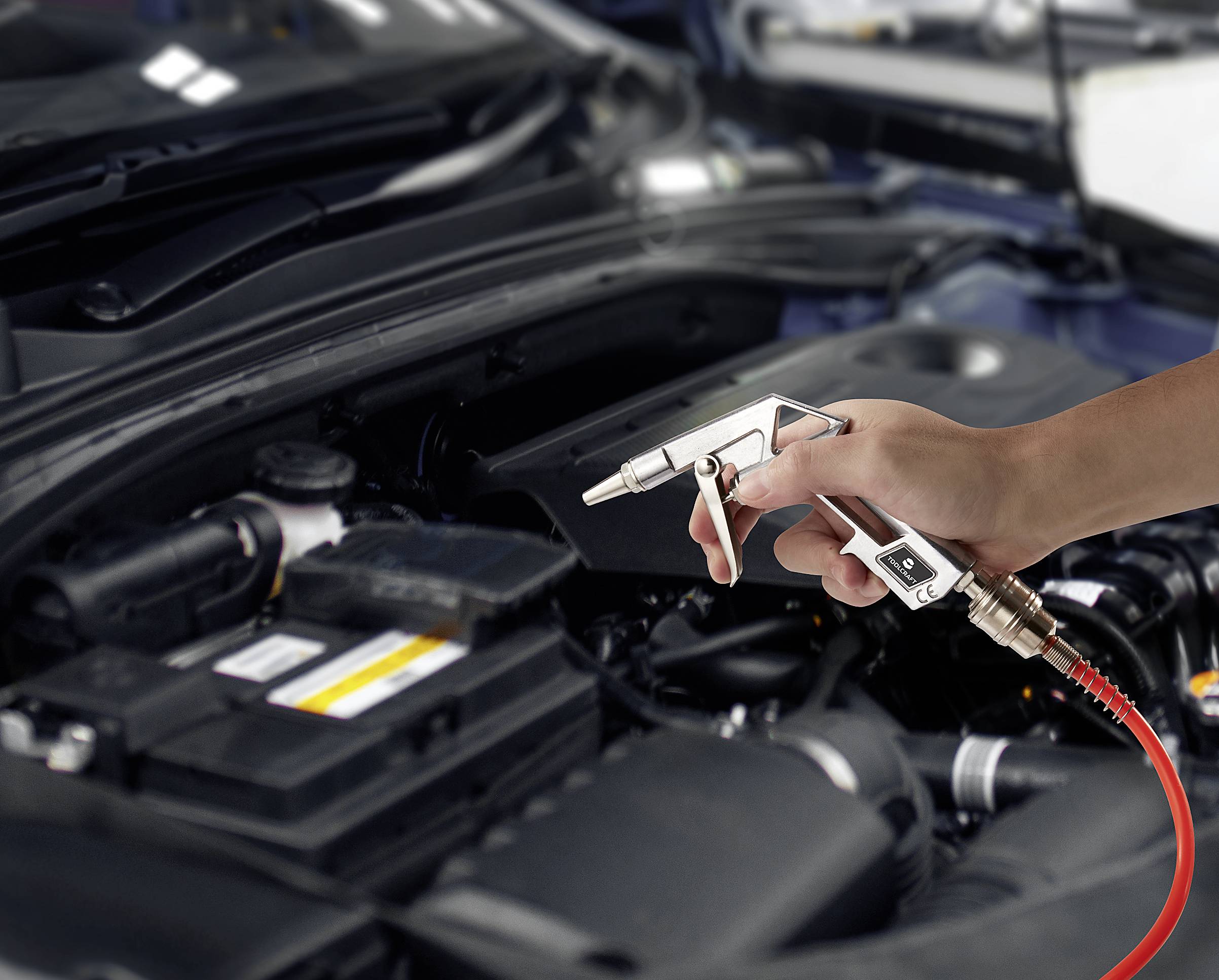 Une main tient un pistolet à air comprimé au-dessus d'un compartiment moteur ouvert. Elle nettoie le moteur en soufflant de l'air sur différentes pièces.