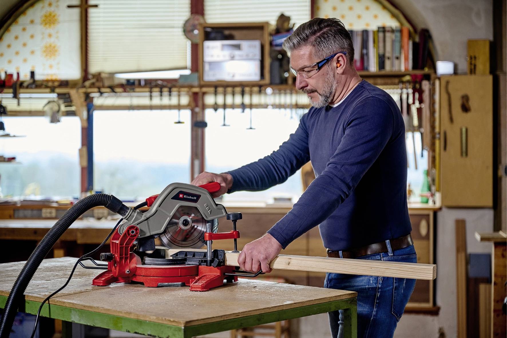 Un homme travaille dans un atelier avec une scie circulaire rouge, en coupant un morceau de bois. Des outils et des étagères sont visibles en arrière-plan.