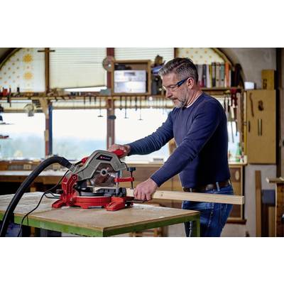 Un homme travaille dans un atelier avec une scie circulaire rouge, en coupant un morceau de bois. Des outils et des étagères sont visibles en arrière-plan.