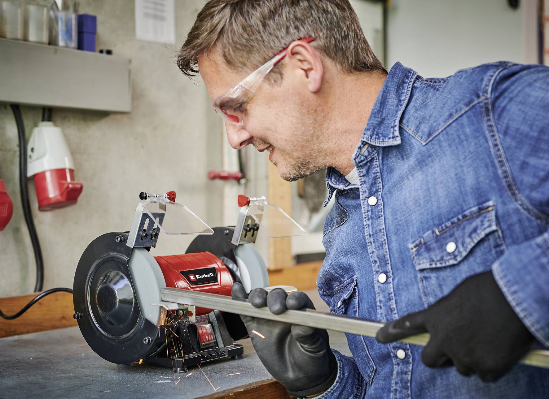 Un homme portant des lunettes de protection et des gants est en train de poncer une barre métallique sur un établi à l'aide d'un outil électrique de meulage.