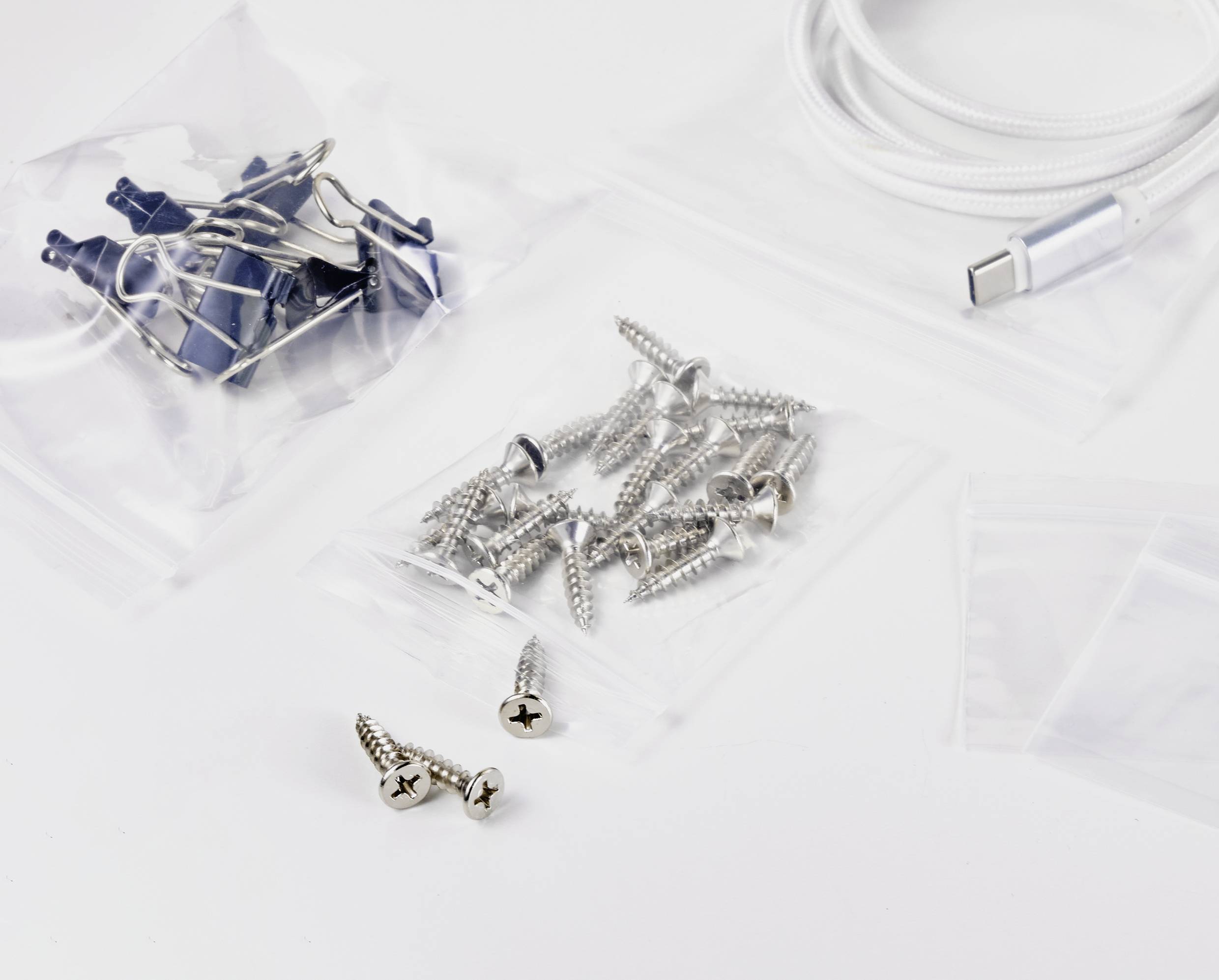 Des pinces de bureau, des vis et un câble de charge organisés dans des sachets transparents sur une surface blanche.