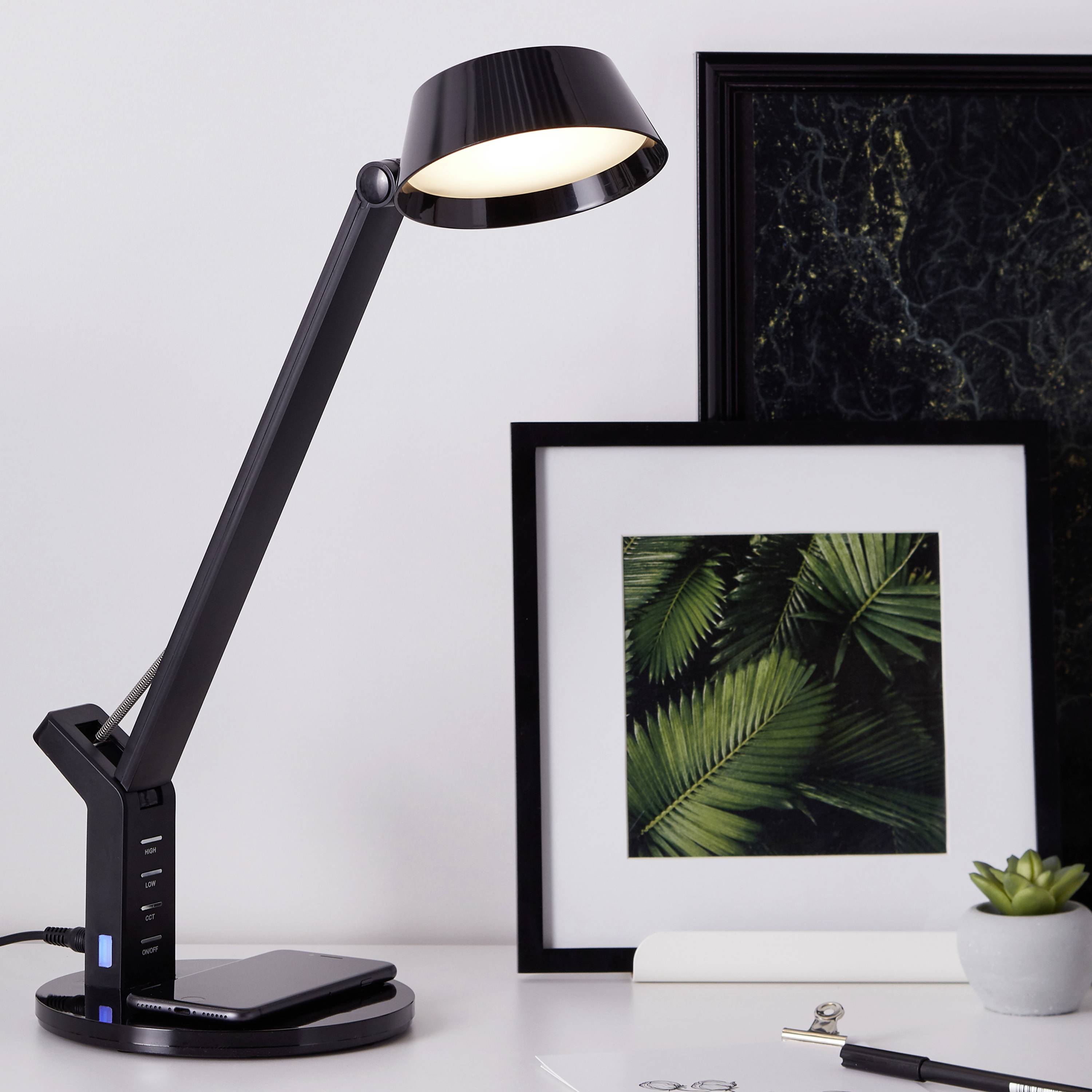 La lampe de bureau éclaire une table sur laquelle se trouvent un cadre photo avec des feuilles vertes et un cahier blanc.