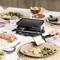 Raclette-grill sur une table dressée avec salade, pommes de terre, jambon et tomates. Une main fait couler du fromage fondu sur l'assiette.