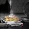 Une pile de pancakes saupoudrés de sucre glace, avec des framboises et des myrtilles sur une assiette. Un gaufrier est posé à côté sur le plan de travail.