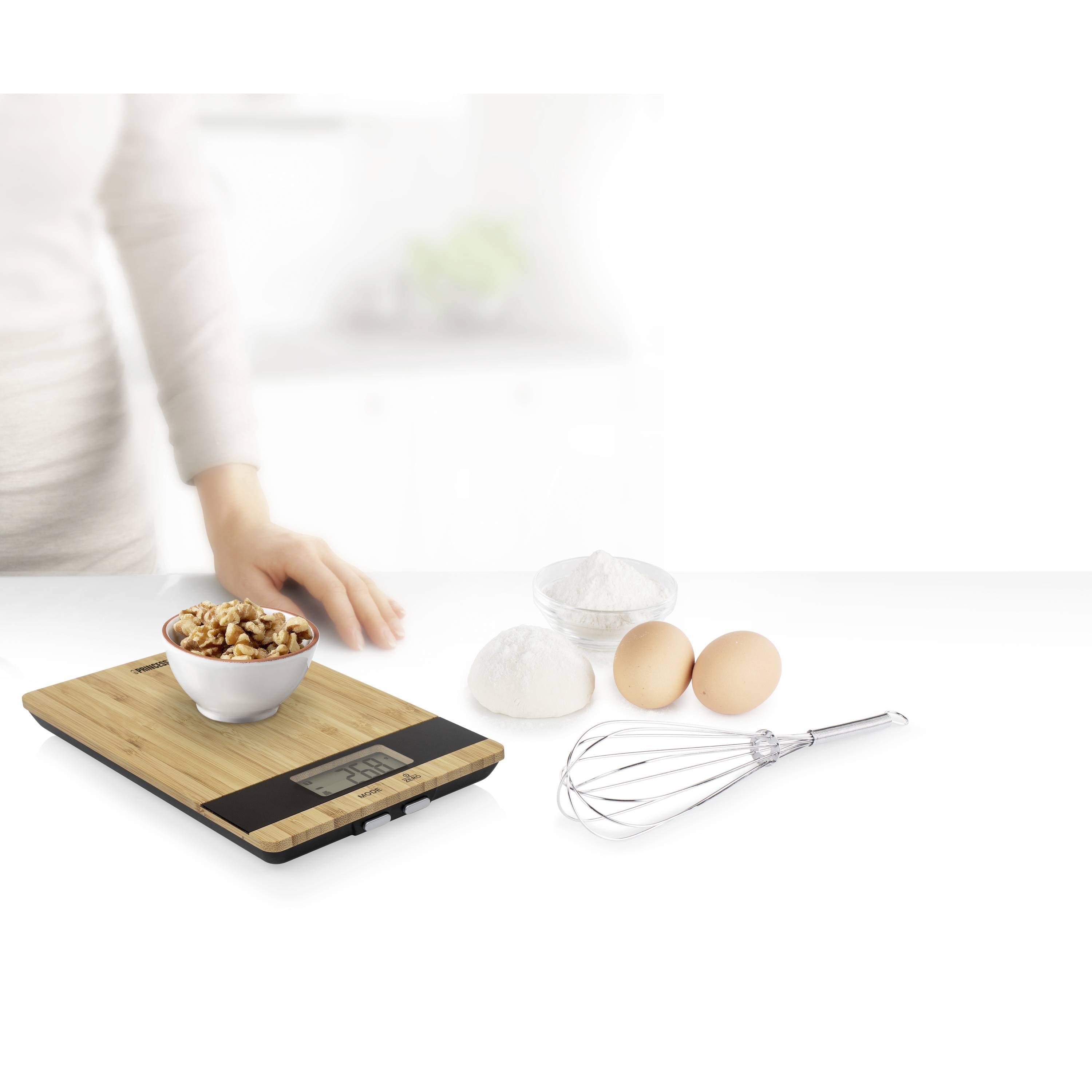 Une femme pèse des ingrédients sur une balance de cuisine numérique : un bol de noix. En arrière-plan, des œufs, de la farine dans des bols, et un fouet.