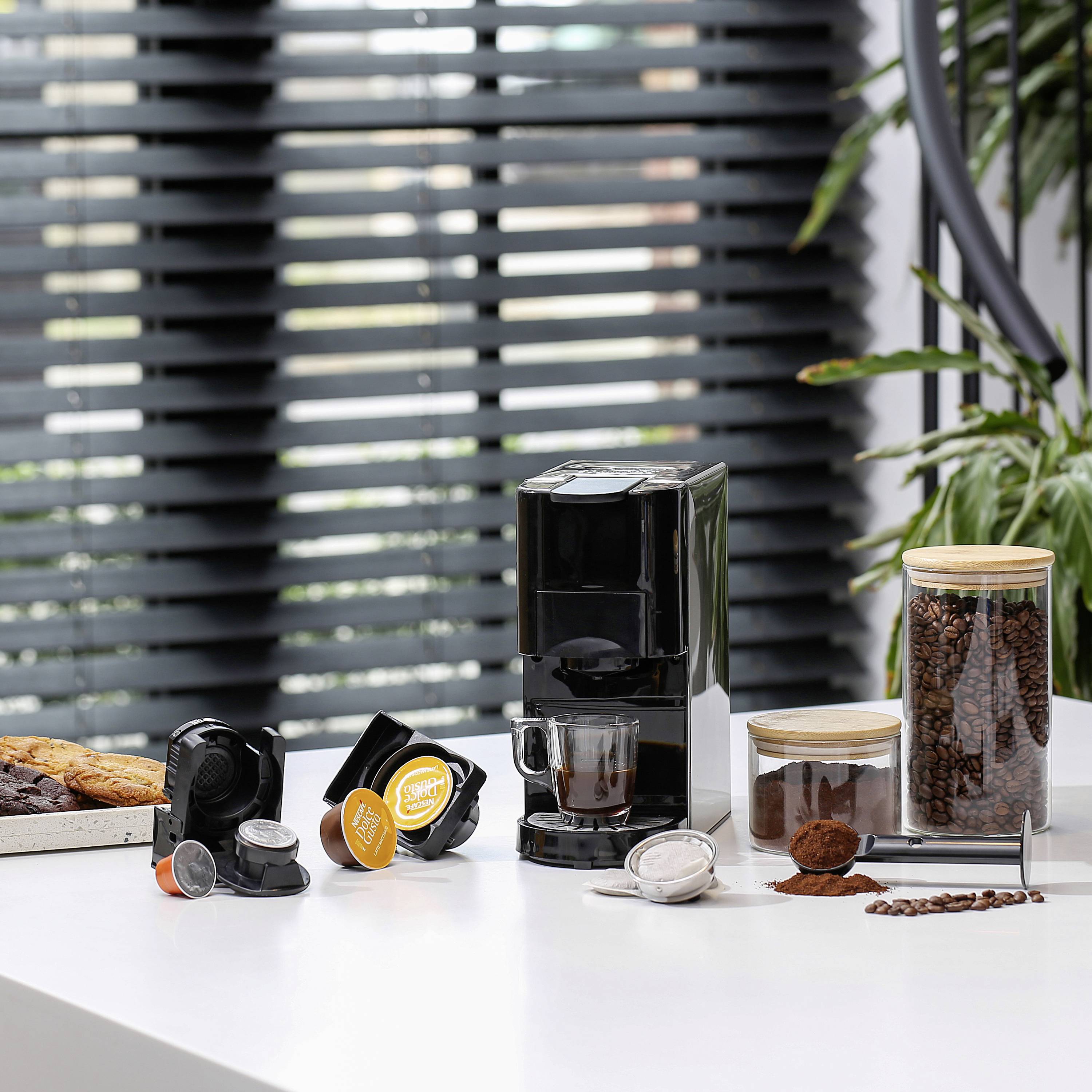 Machine à café avec des capsules et des grains de café au premier plan, de grandes fenêtres avec des plantes en arrière-plan, illustrant le plaisir de déguster du café.