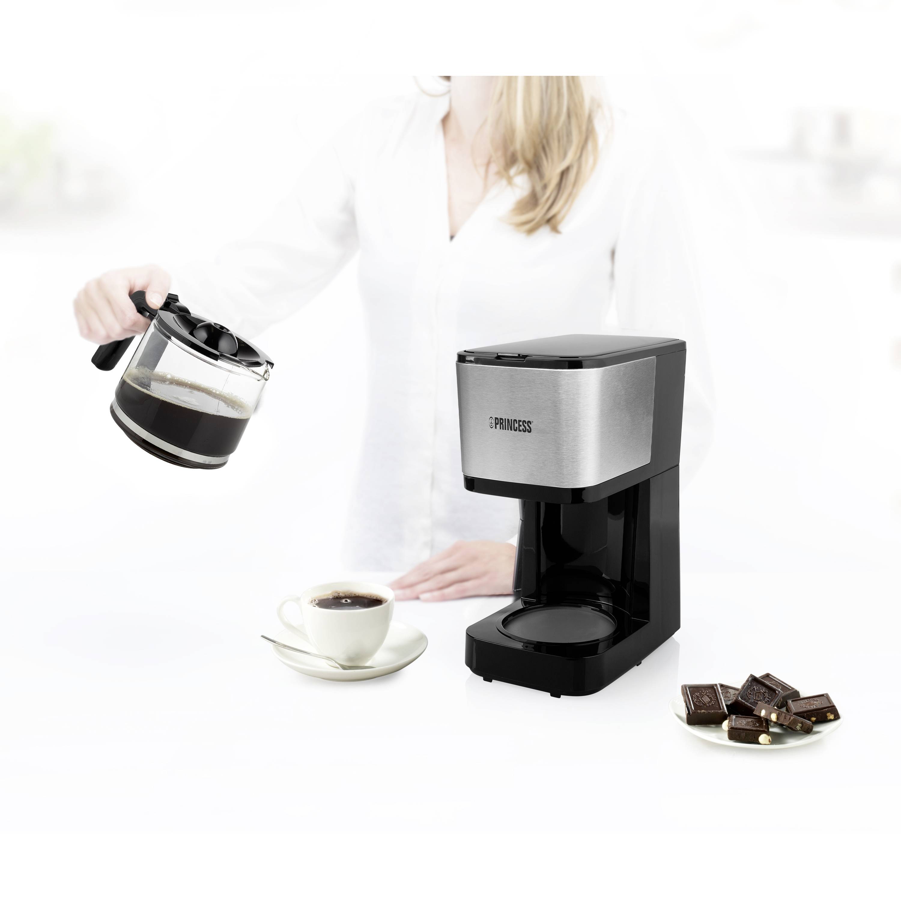 Une femme verse du café d'une cafetière dans une tasse à côté d'une machine à café. Un cuillère et quelques morceaux de chocolat sont posés à proximité.