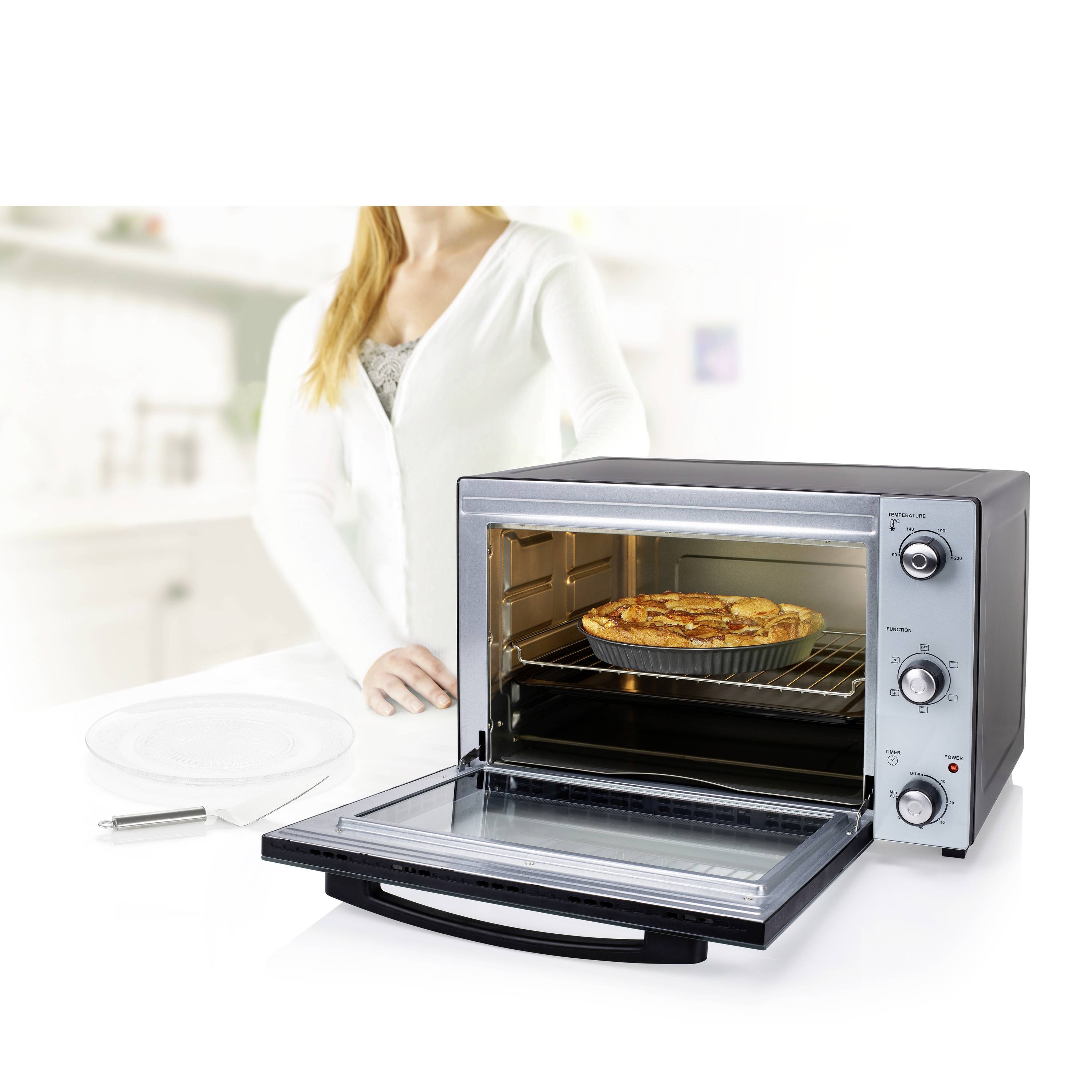 Une femme se tient dans une cuisine à côté d'un four de table ouvert contenant un gâteau fraîchement cuit. Le four est propre et moderne.