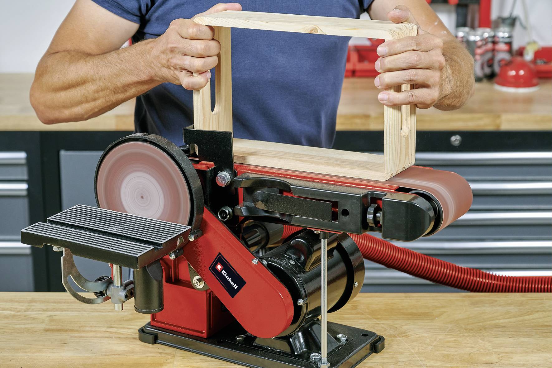 Une personne utilise une ponceuse à bande pour travailler une pièce de bois rectangulaire. La bande de ponçage et la table de travail sont visibles.