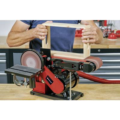 Une personne utilise une ponceuse à bande pour travailler une pièce de bois rectangulaire. La bande de ponçage et la table de travail sont visibles.