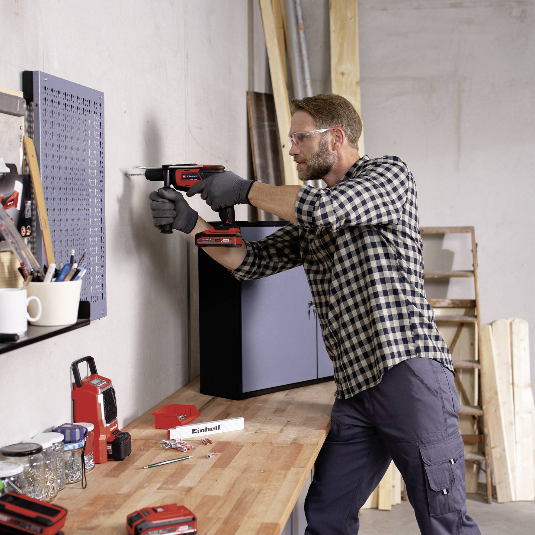 Un homme utilise une perceuse pour faire un trou dans un mur. À côté de lui, on peut voir une table avec des outils et une échelle.
