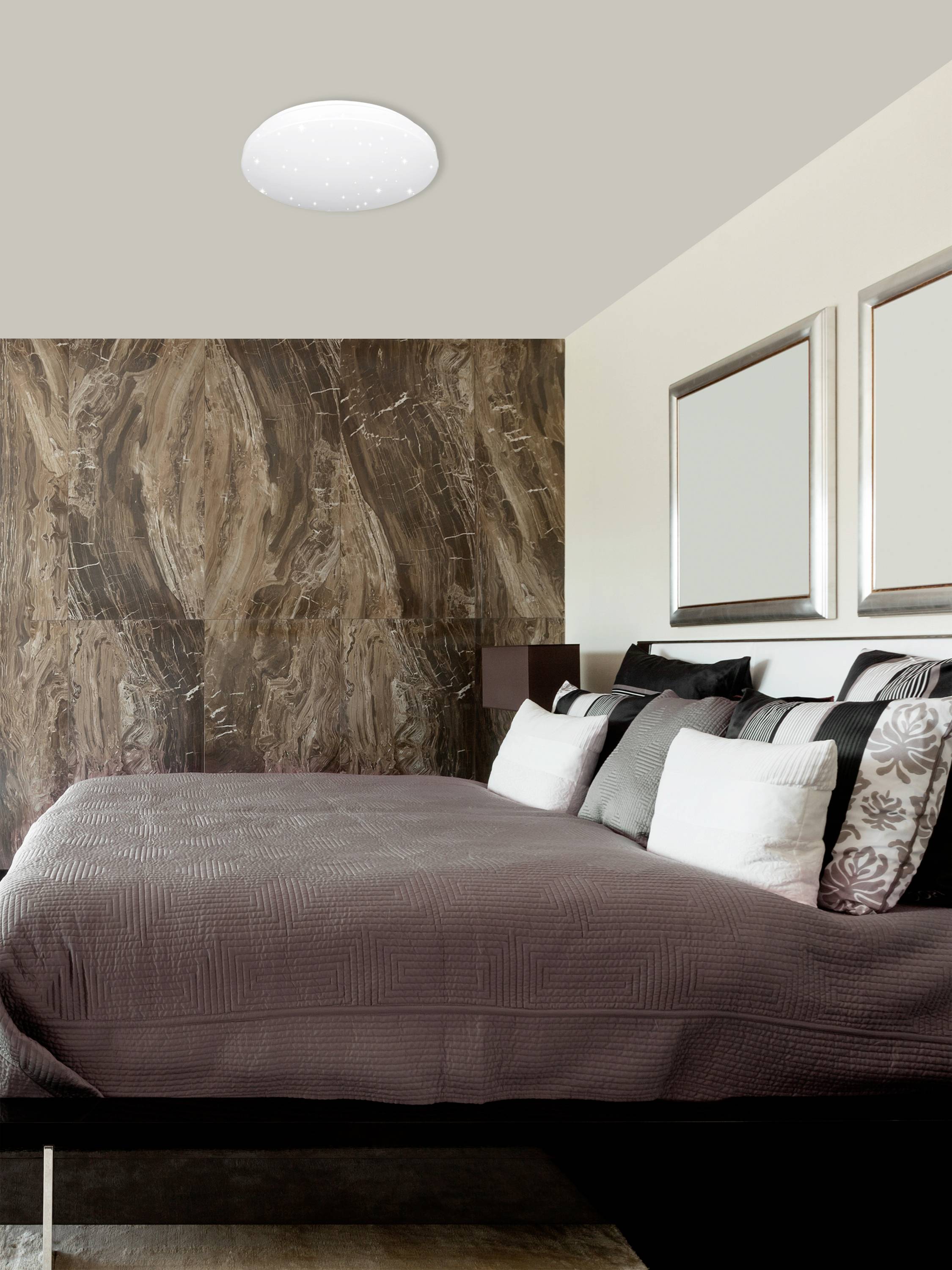 Chambre à coucher avec un grand lit, décoré de plusieurs coussins. Au plafond se trouve une lumière ronde et moderne.