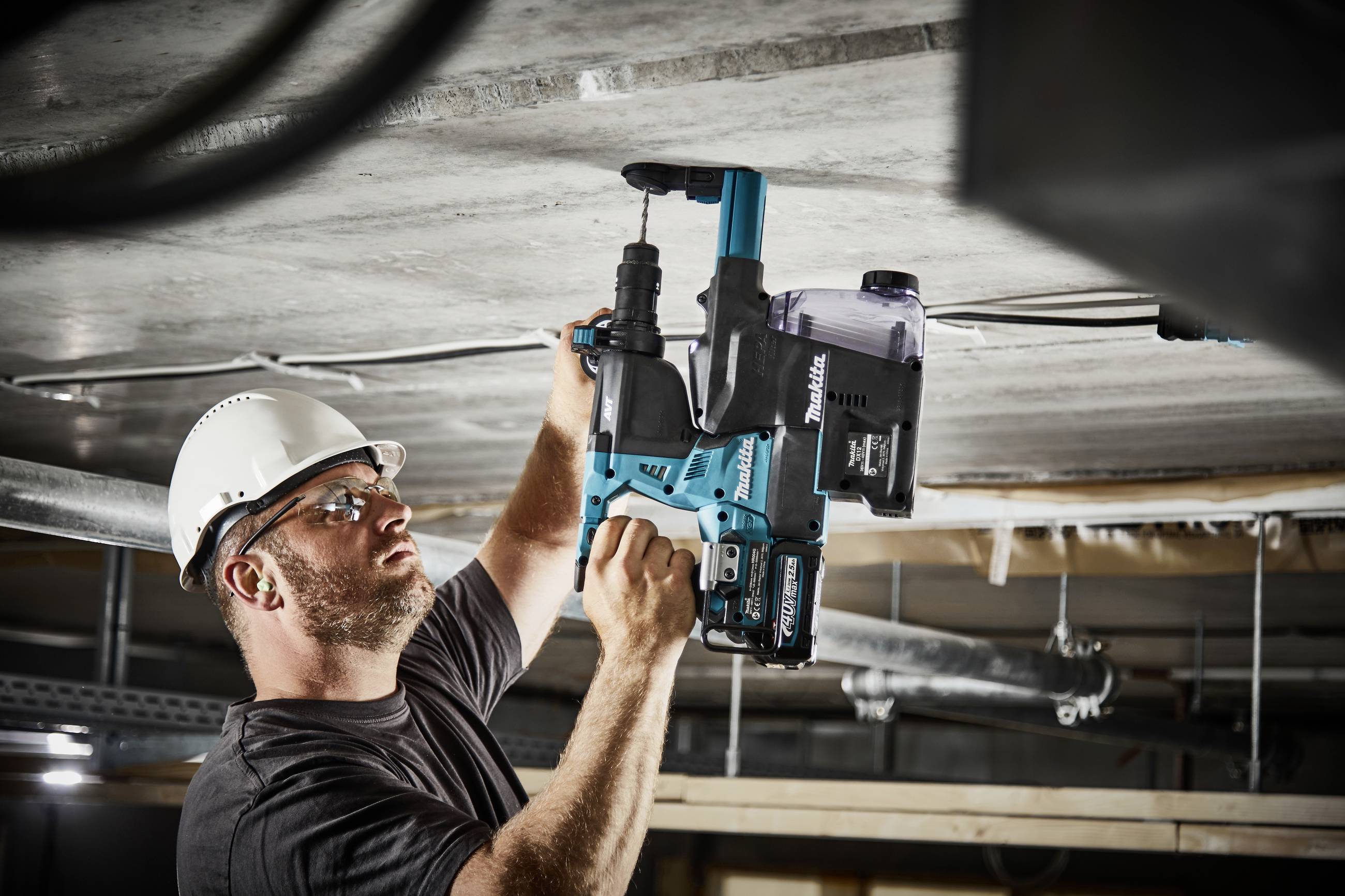 Un ouvrier en tenue de protection utilise un outil électrique bleu pour percer une surface en béton, probablement au plafond.