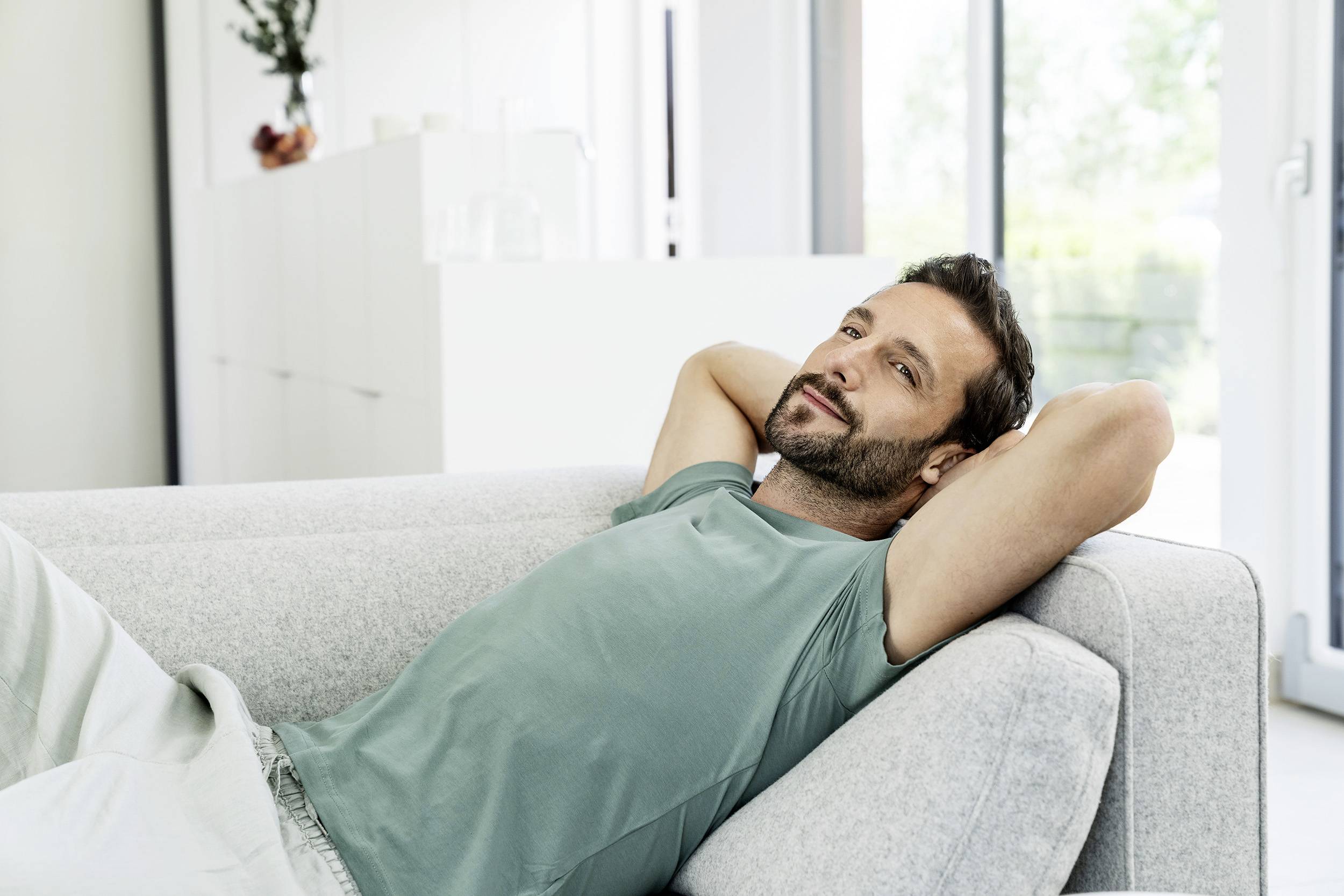 Un homme est allongé de manière détendue sur un canapé, les mains derrière la tête. Il porte un t-shirt clair et sourit en regardant sur le côté. À l'arrière-plan, on aperçoit des meubles clairs et une fenêtre.