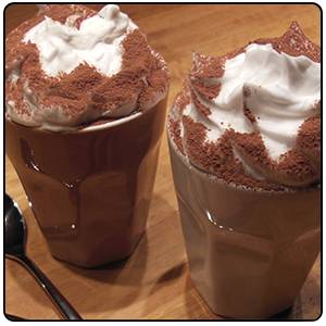Deux tasses de chocolat chaud surmontées de crème chantilly, posées sur une table en bois. Une cuillère est placée à côté.