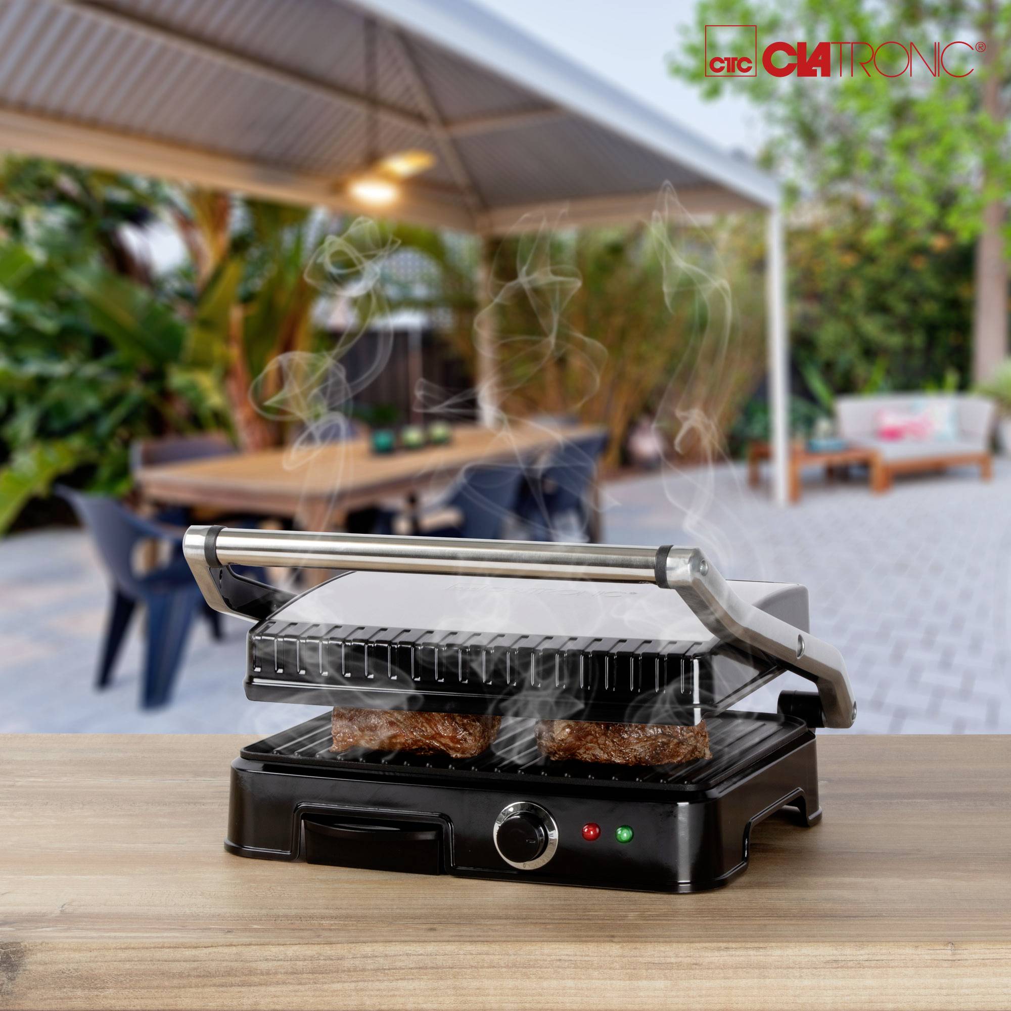 Un grill de table fait griller deux sandwichs d'où s'élève de la vapeur. En arrière-plan, on aperçoit une terrasse couverte avec des meubles de jardin.