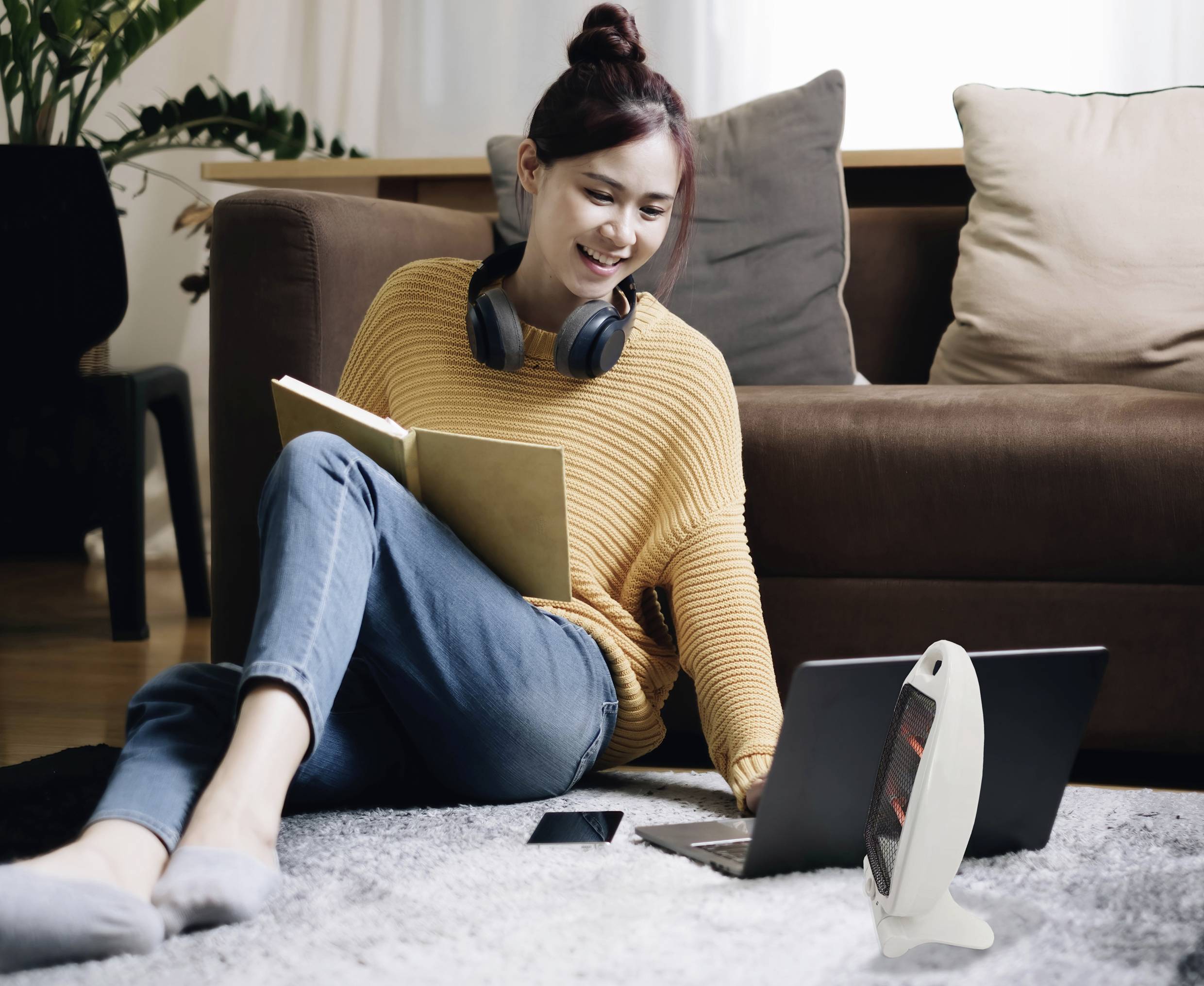 Une femme est assise sur un tapis, portant des écouteurs et lisant un livre. Un ordinateur portable est posé devant elle, et un canapé se trouve en arrière-plan.