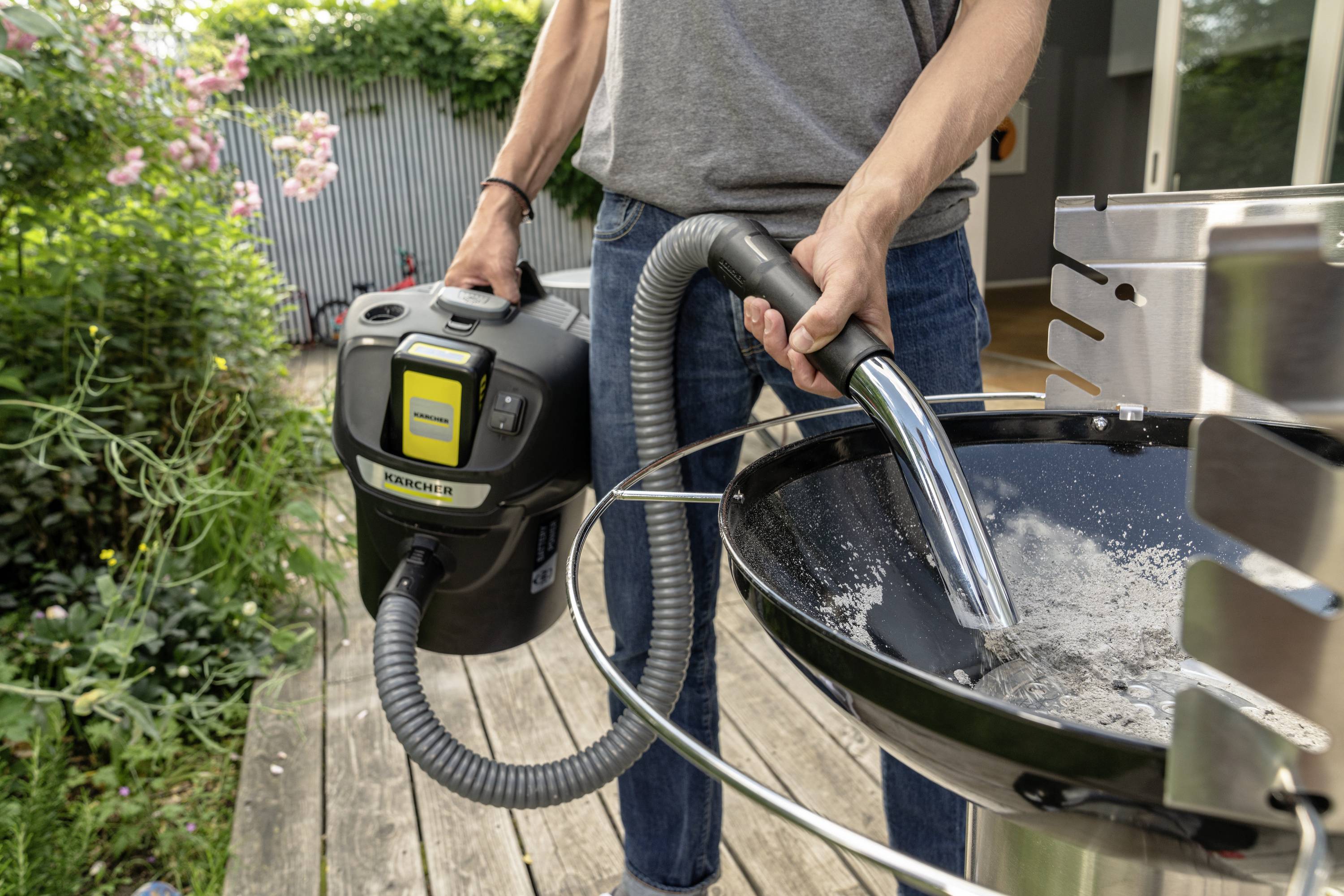 Une personne aspire des cendres d'un grill à l'aide d'un aspirateur à cendres portable. Des plantes sont visibles en arrière-plan.