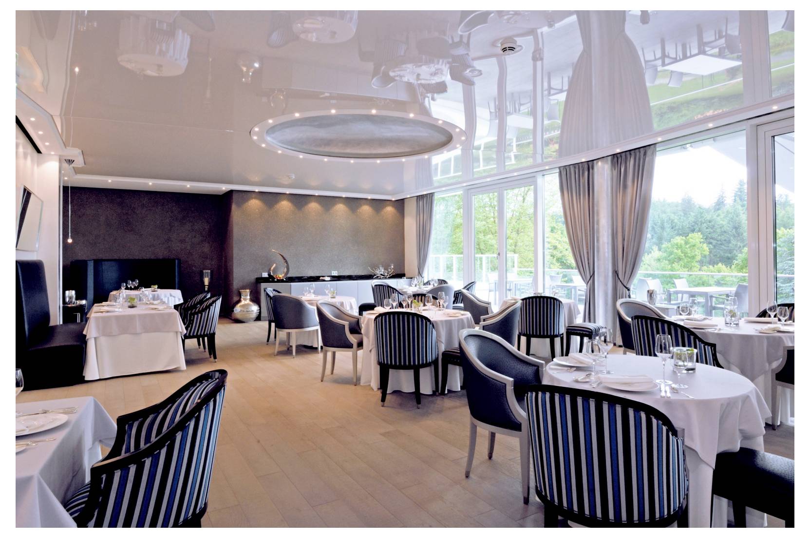 Salle de restaurant élégante avec des tables dressées, des chaises rayées et de grandes fenêtres donnant sur la verdure.