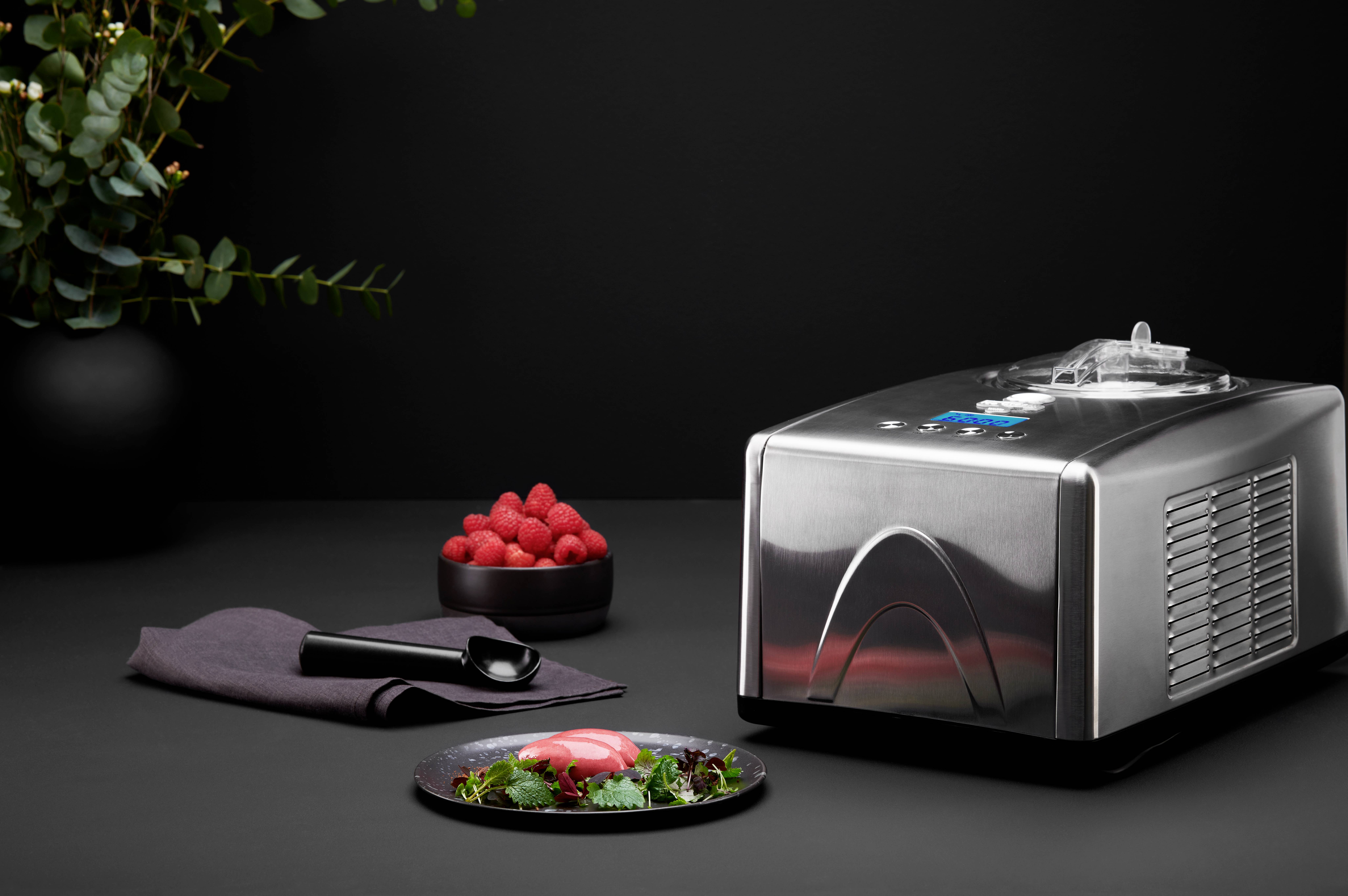 Une machine à glace chromée à côté d'une assiette de dessert, d'un bol de framboises et d'une cuillère noire, sur un fond sombre.