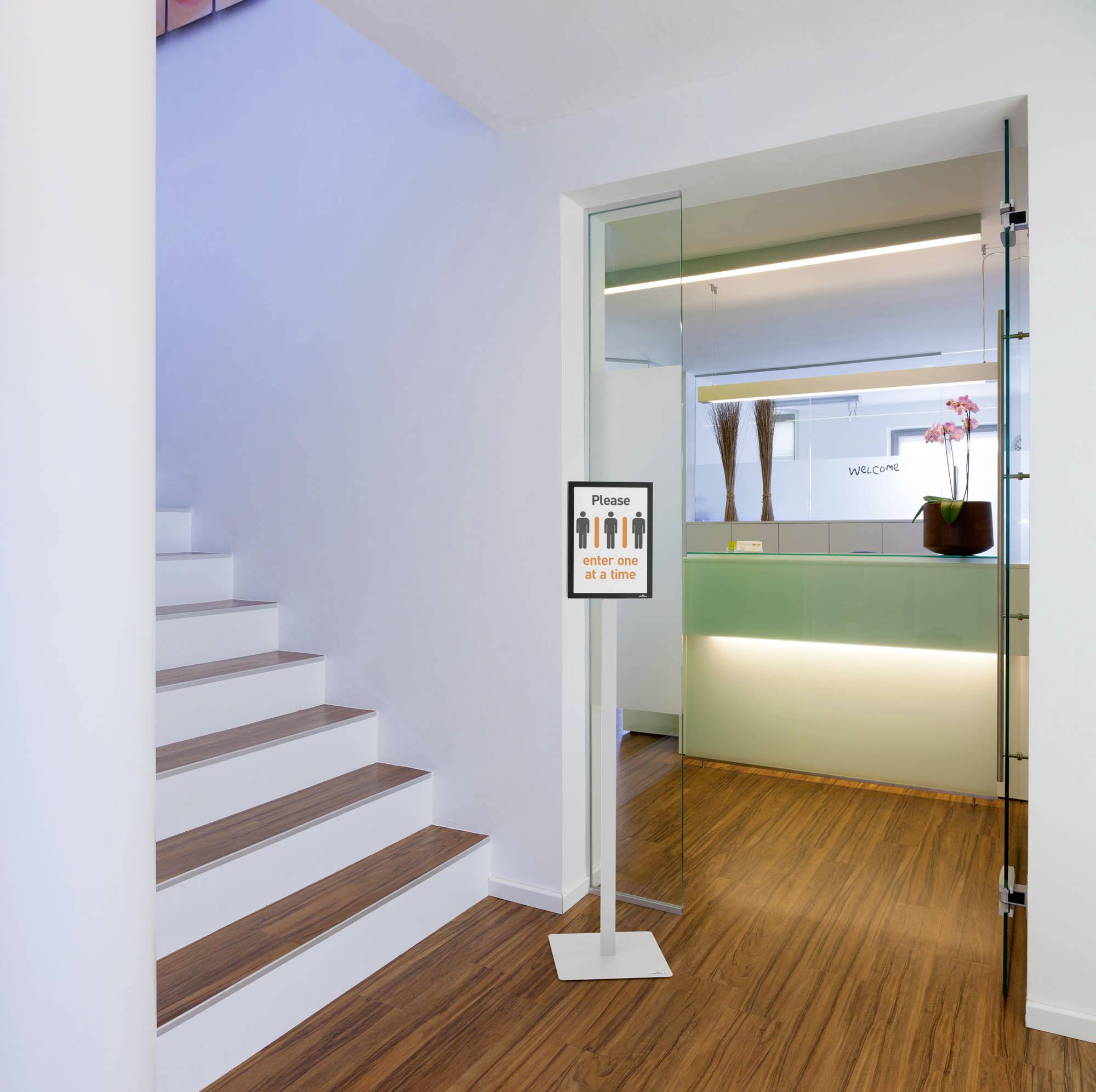 Une porte vitrée ouverte à côté d'un escalier, avec un panneau indiquant 'Veuillez garder vos distances'. Derrière, un espace d'accueil moderne.