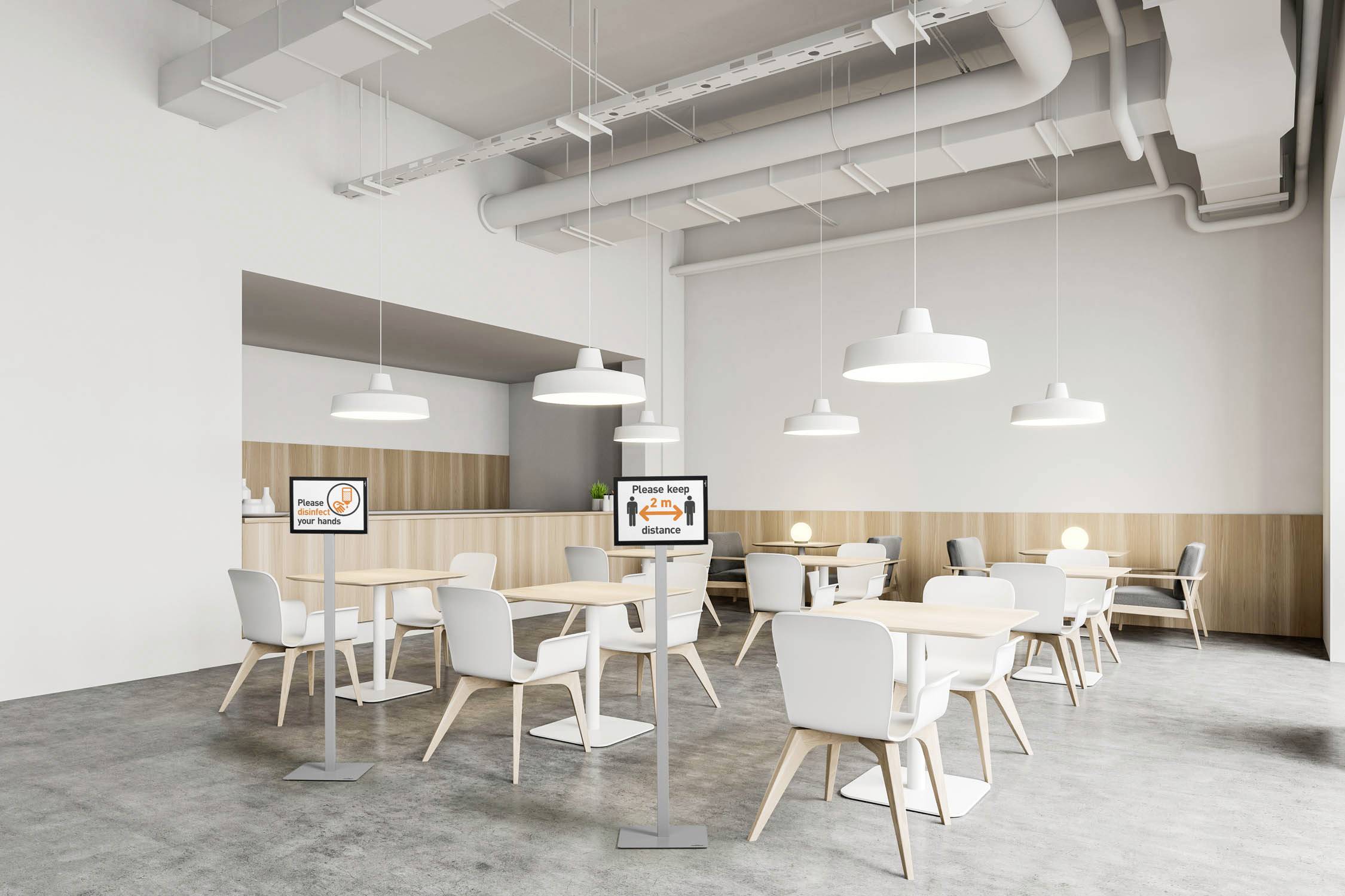 Une salle à manger moderne avec des tables et des chaises blanches, des suspensions lumineuses et une décoration neutre. Deux écrans affichent des informations de sécurité.